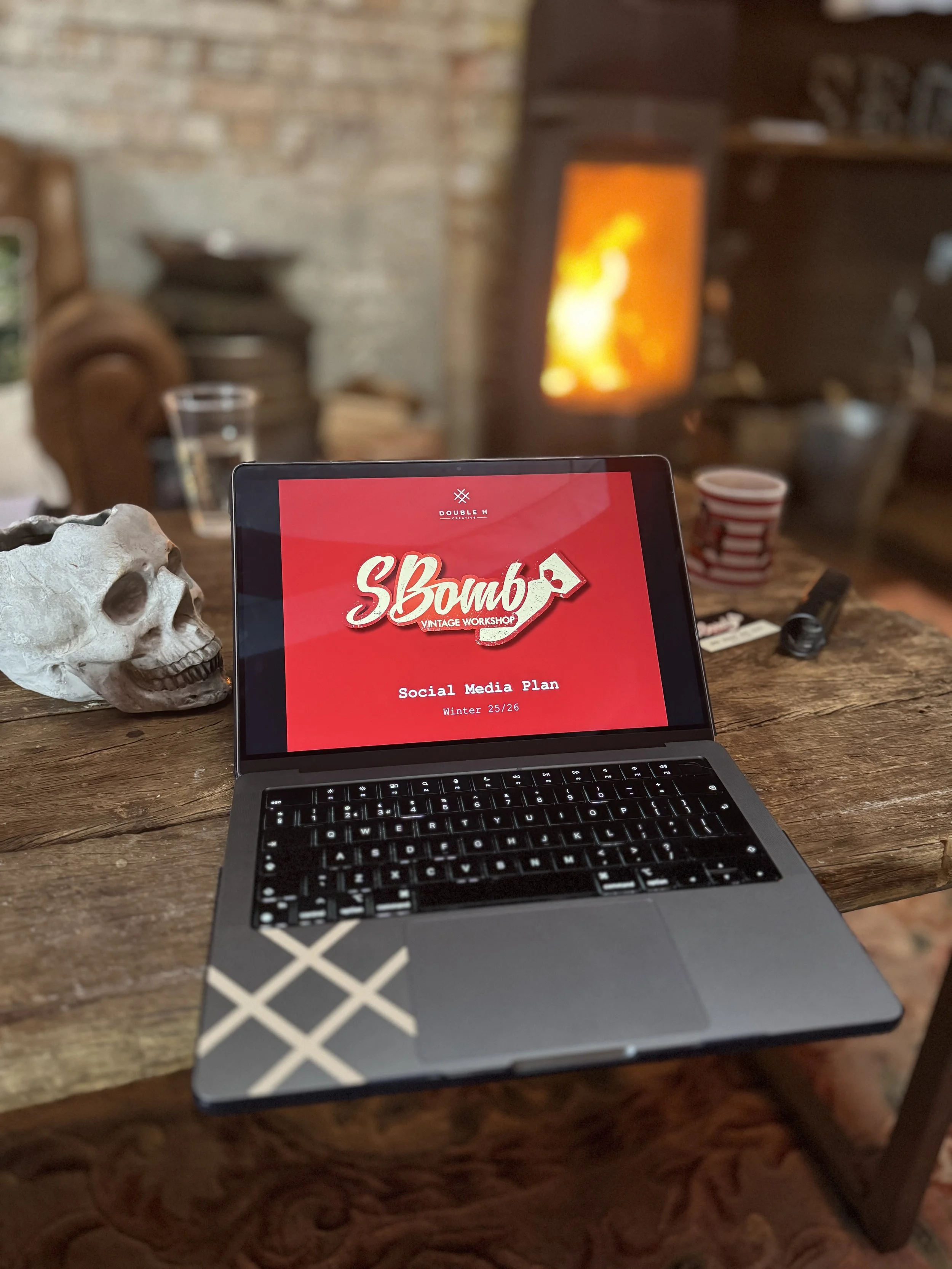A laptop on a wooden table displaying a red screen with the text 'S Bomb Vintage Workshop' and 'Social Media Plan Winter 25/26'. A skull sculpture is on the left side of the table, and a glass and striped cup are in the background. A blurry fireplace is visible in the background.