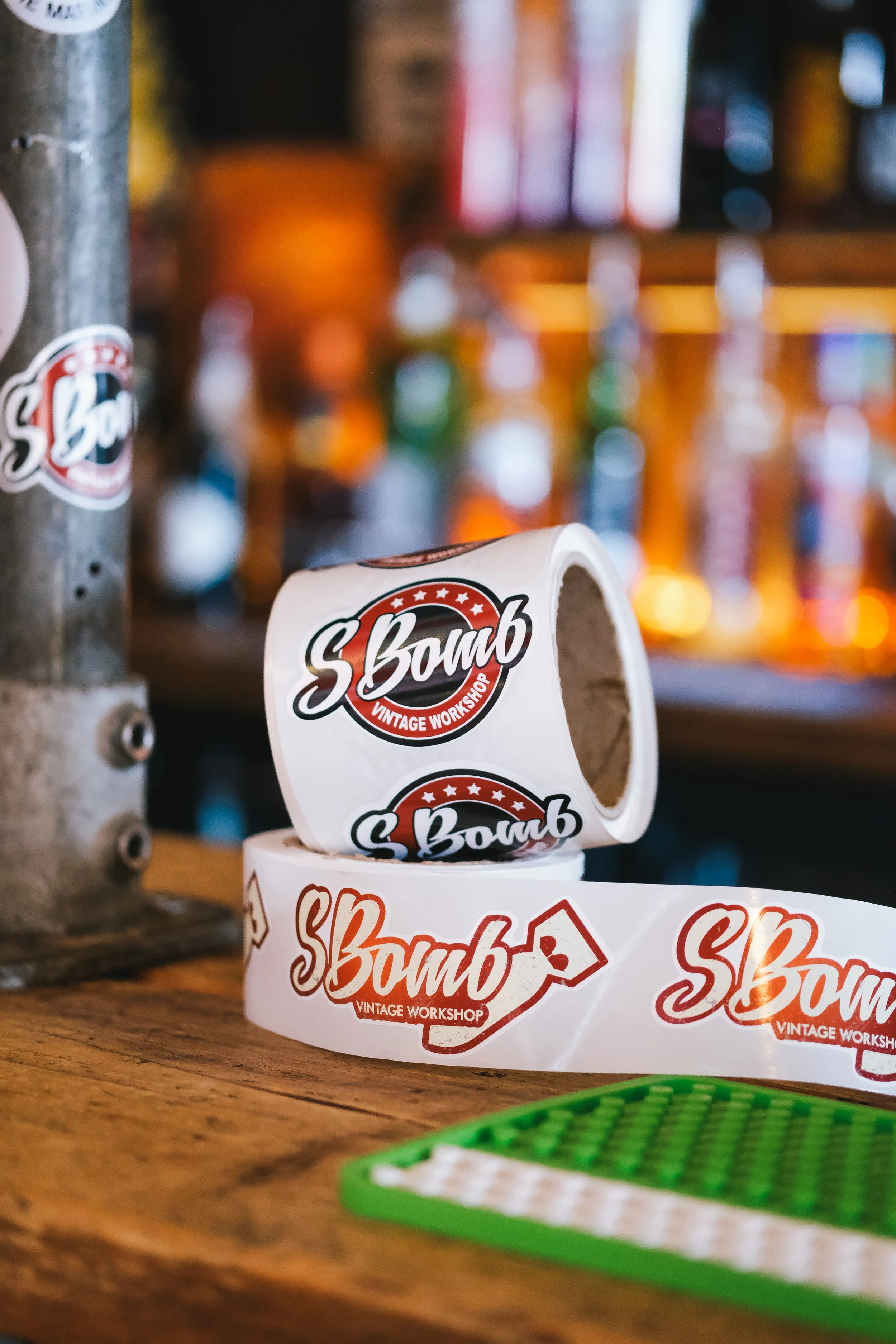 Close-up of a roll of S.Bomb vintage workshop stickers on a wooden surface in a bar or brewery setting, with a blurred background of bottles and colorful lights.