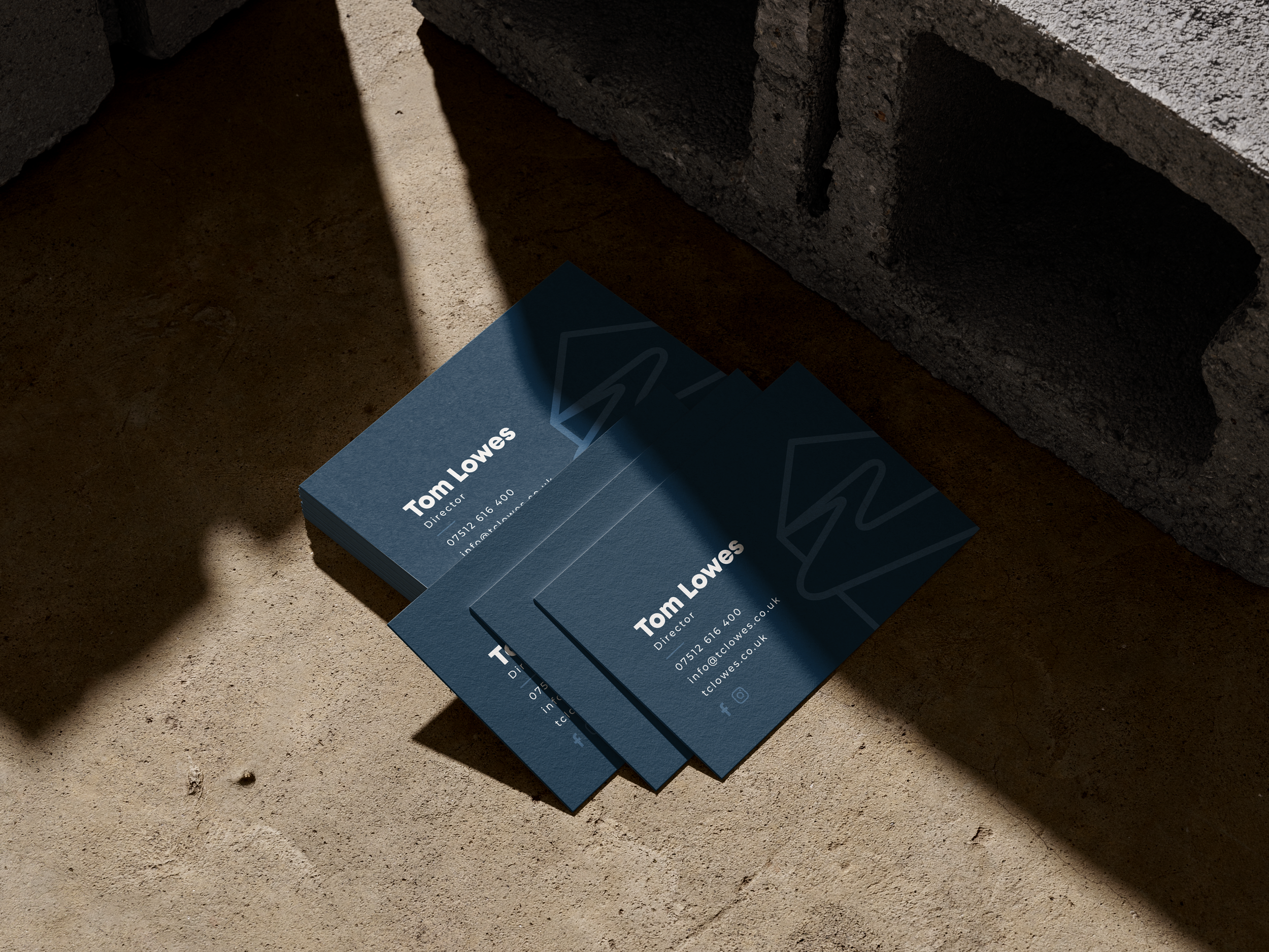 Stack of blue business cards with contact information for Tom Lowes, a director, placed on a concrete surface next to a brick wall, with shadows cast across the scene.