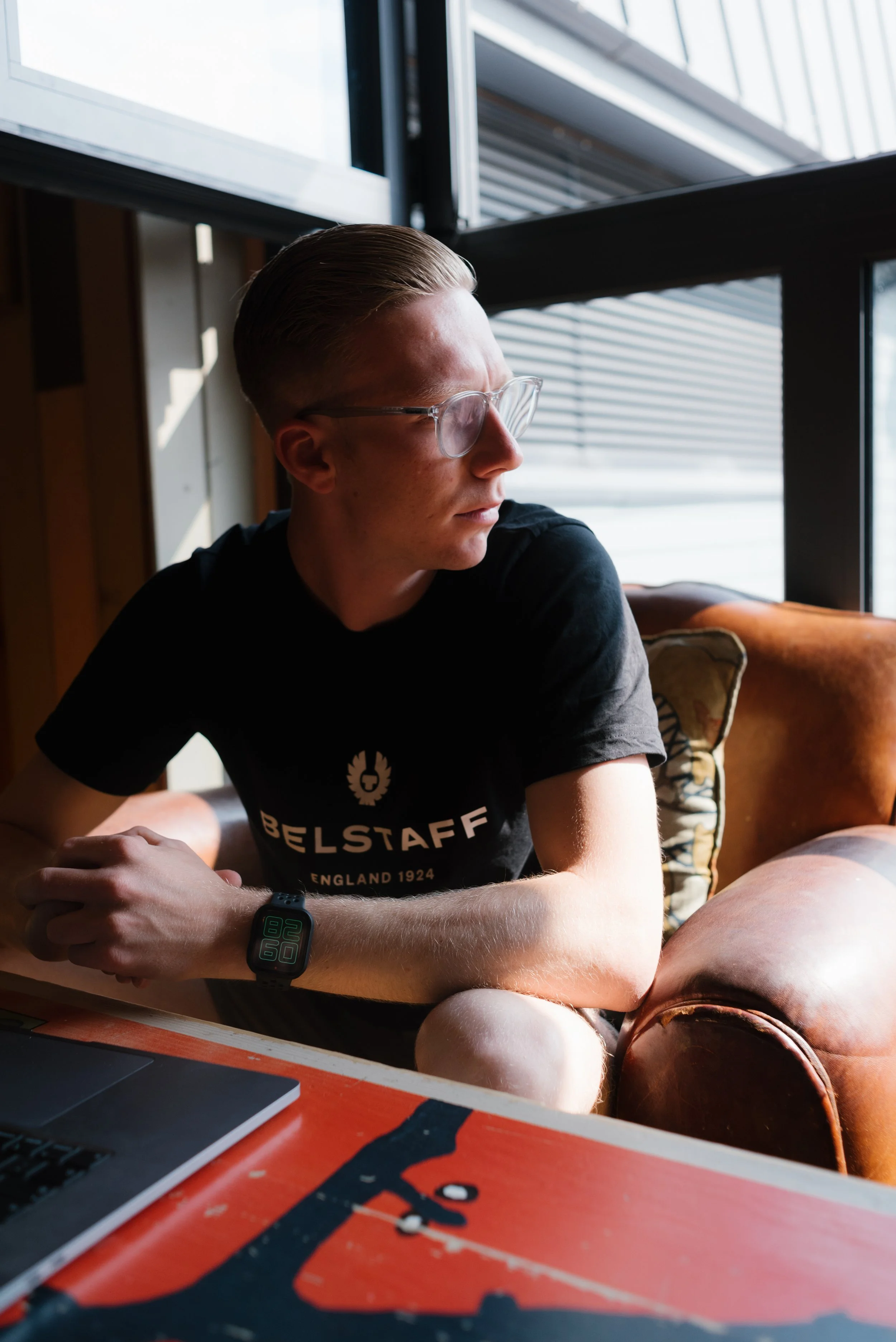 A young man with glasses and short, styled hair, wearing a black t-shirt with 'BELSTAFF' written on it, sitting by a large window with sunlight. He has a smartwatch on his left wrist and is looking to his right. There is a laptop on a table in the foreground and a leather armchair behind him.