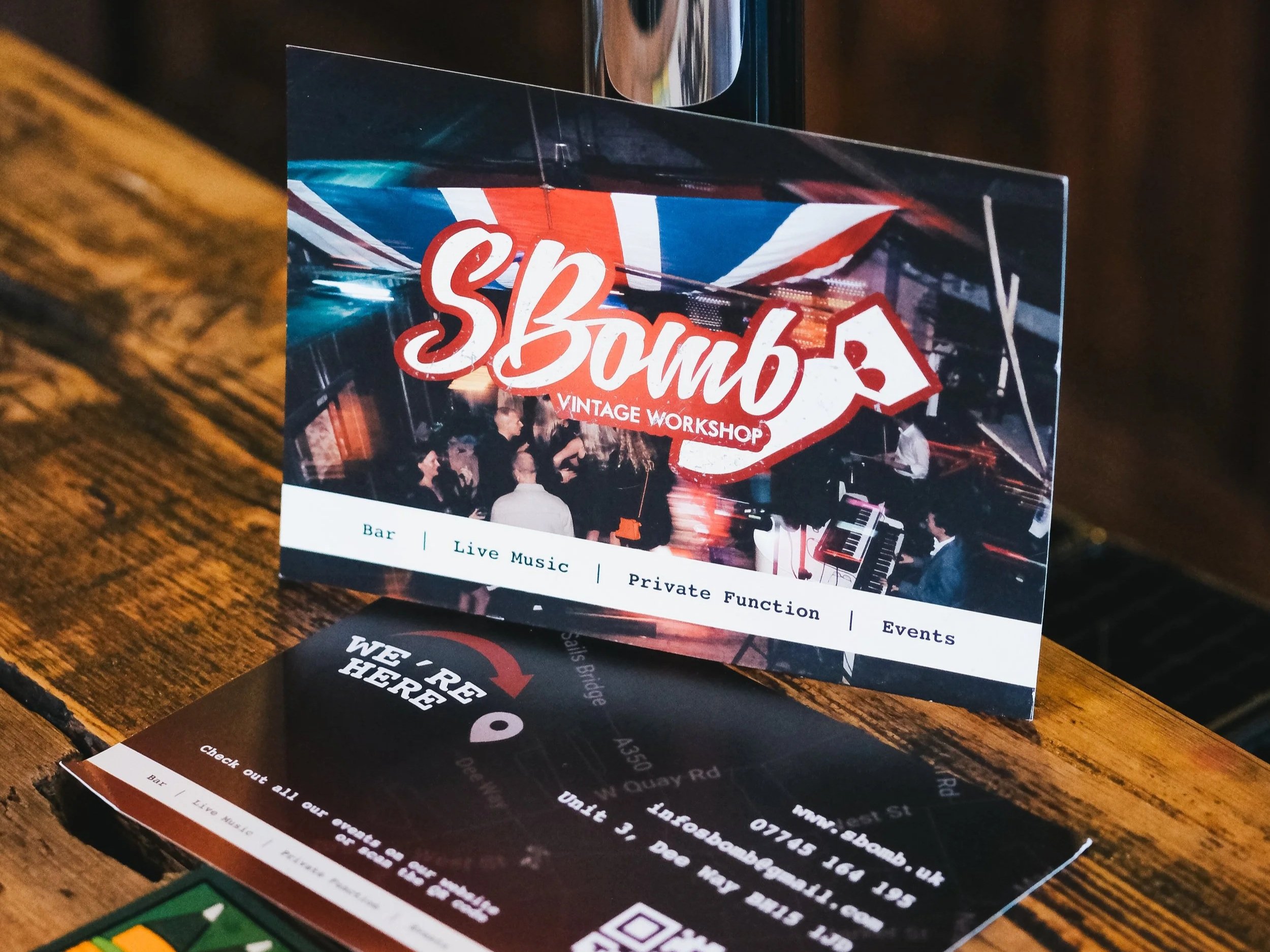 A stack of event flyers on a wooden table, announcing a vintage workshop called 'SBomb' with live music, private functions, and events, featuring a colorful image of a lively scene with people and a piano.