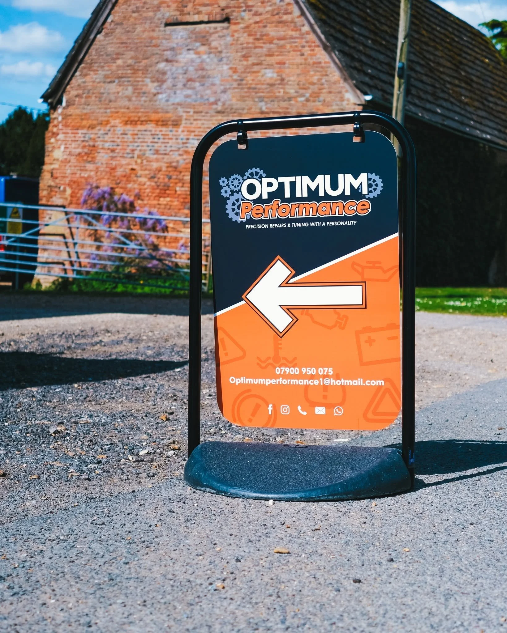 A sign for Optimum Performance with a white arrow pointing left, placed on a wheeled stand outside near a brick building and a low fence with purple flowers.