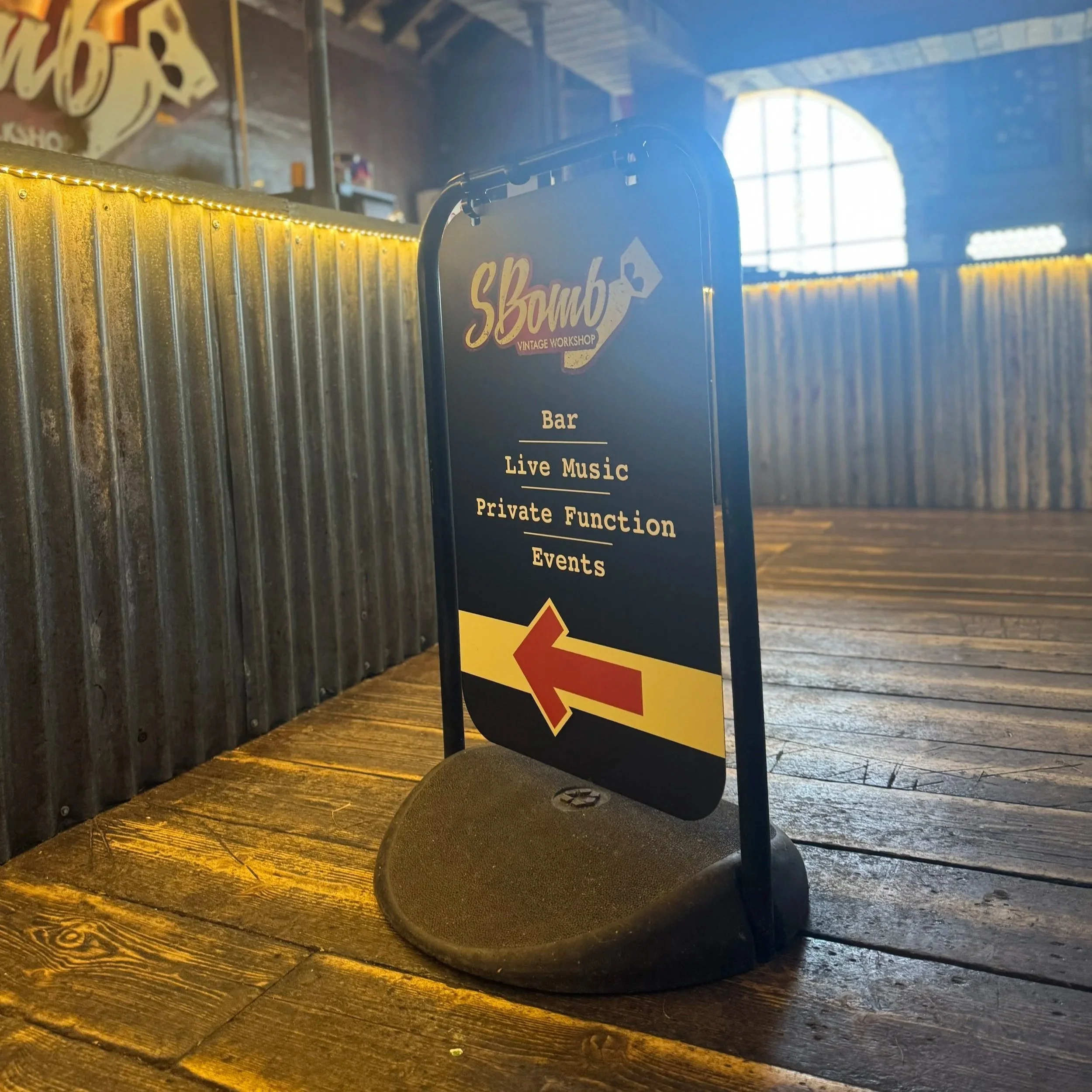 A black signboard with yellow and white text promoting a vintage workshop, bar, live music, events, and private functions, placed on a wooden floor with a rustic wall and large window in the background.