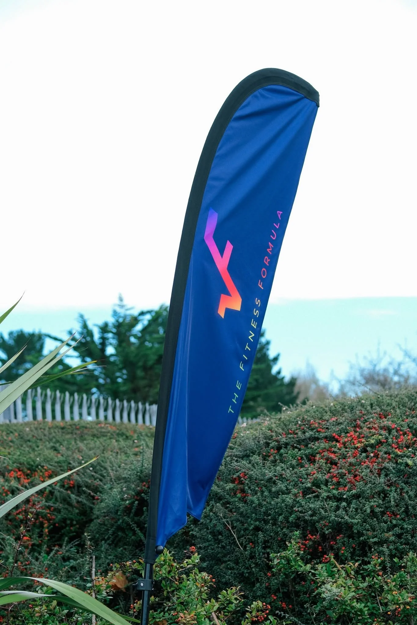 A blue vertical flag with the words 'FITNESS FOR LIFE' in pink and purple, located outdoors among bushes and trees, with a fence and a building in the background.