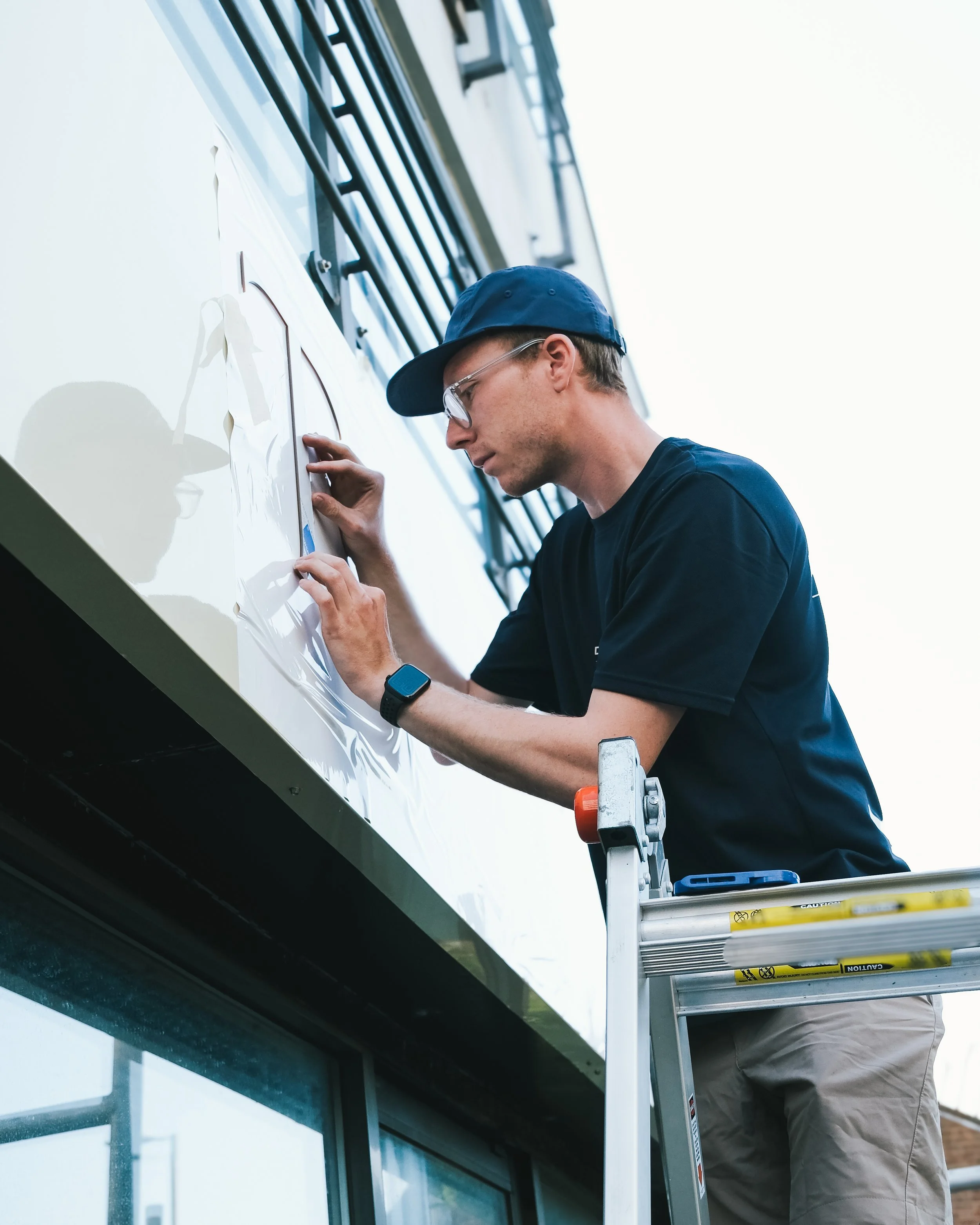 Man working on exterior building signage on a ladder, wearing safety glasses and a baseball cap.