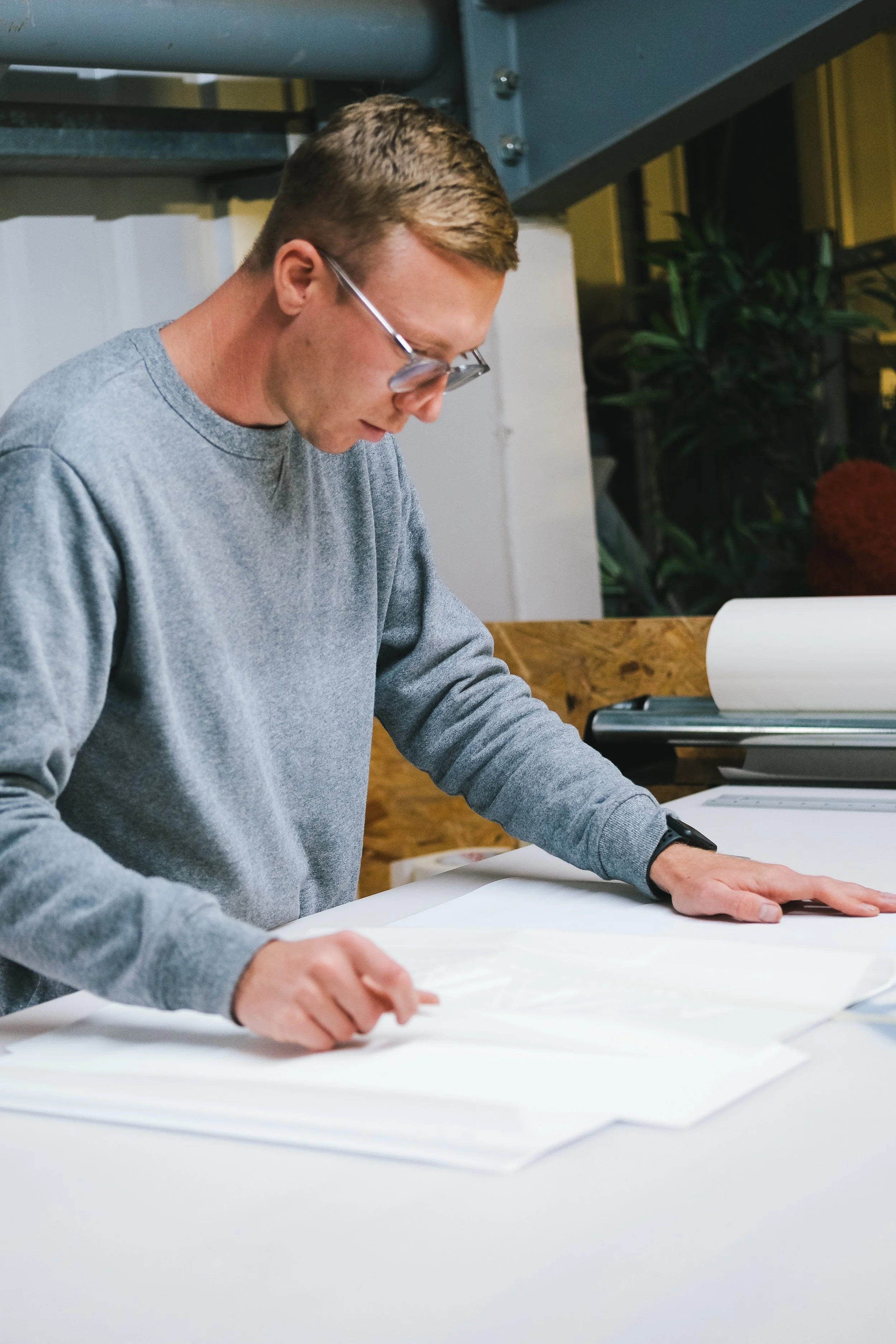 A young man with short hair, glasses, and wearing a gray sweatshirt, standing at a table, looking down at a large sheet of paper or blueprint. There are papers and a laptop on the table, with a background of plants and an industrial-style setting.