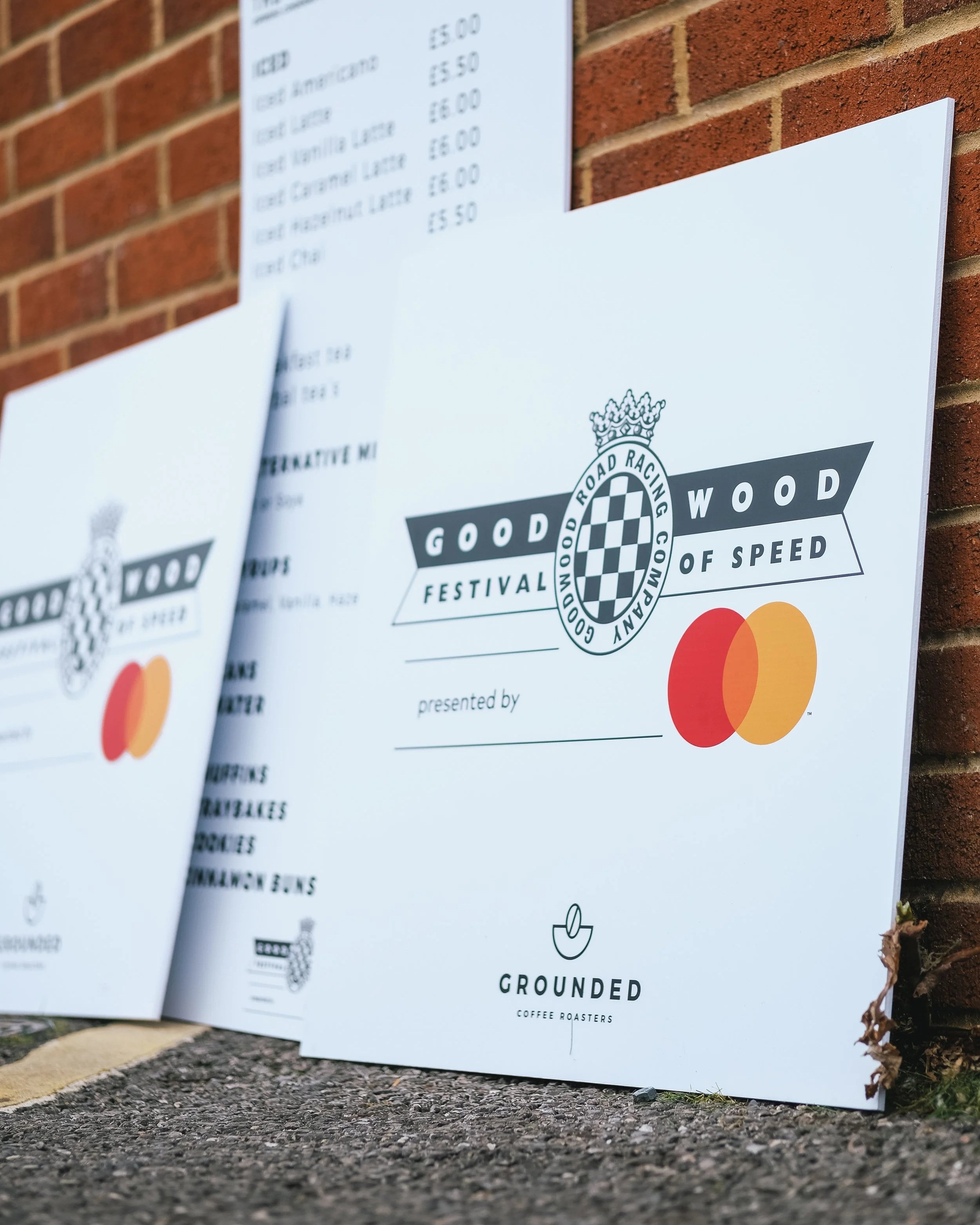 Outdoor promotional signs with event details and logos, leaning against a brick wall, advertising a festival called 'Good Wood of Speed' featuring coffee roasters, sponsored by Mastercard. The signs are on a paved surface, with dried leaves at the bottom right corner.
