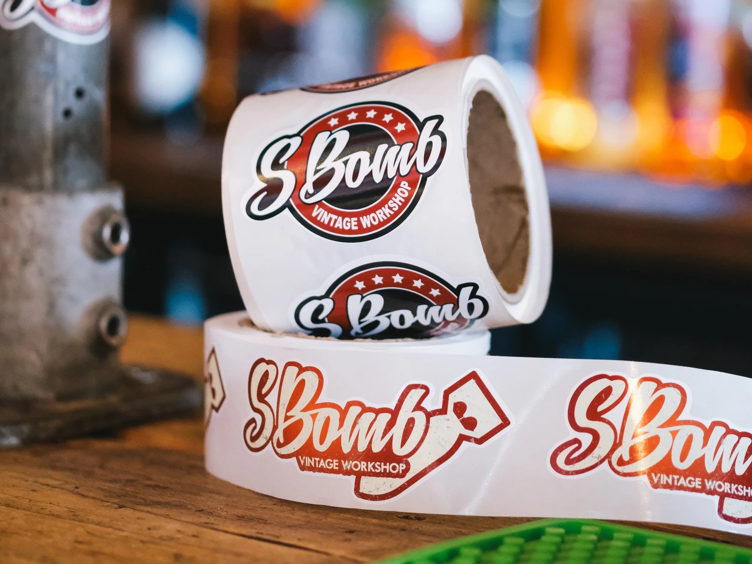 Close-up of a roll of tape with a logo reading 'S Bomb Vintage Workshop' in a red, white, and black design. The tape is on a wooden surface with blurred objects in the background.