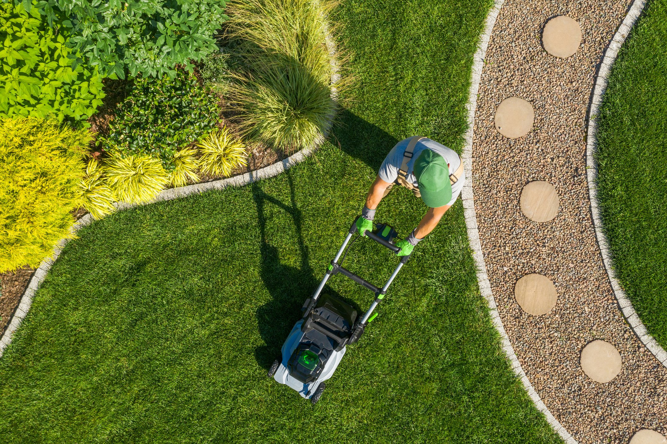 A person mowing a lush green lawn with a push lawn mower on a curved garden path surrounded by vibrant plants and shrubs.