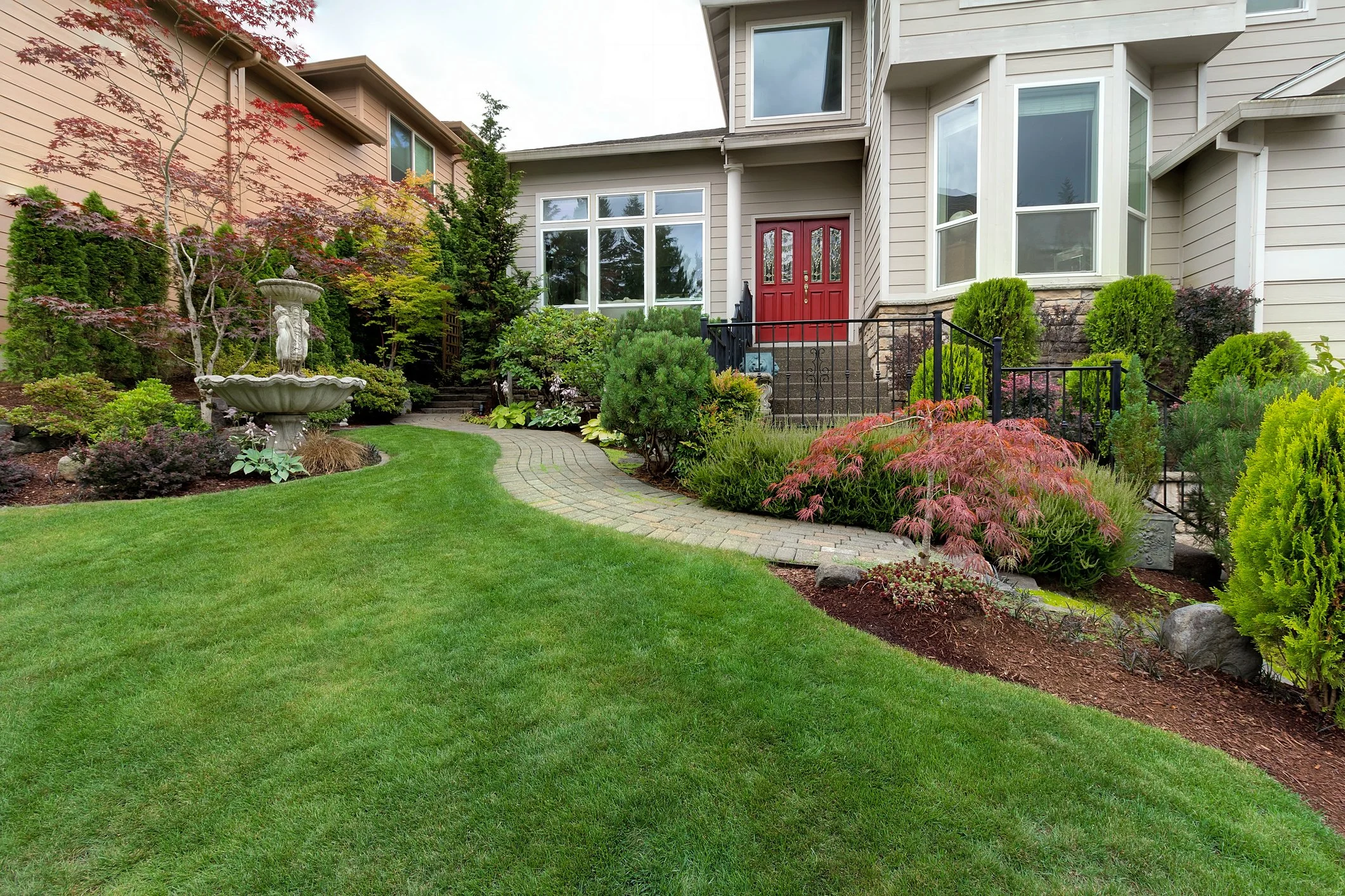 Well-maintained front yard of a house with a brick pathway, lush green grass, and diverse bushes and trees, including a Japanese maple. The house has a red front door, large windows, and a decorative fountain on the lawn.