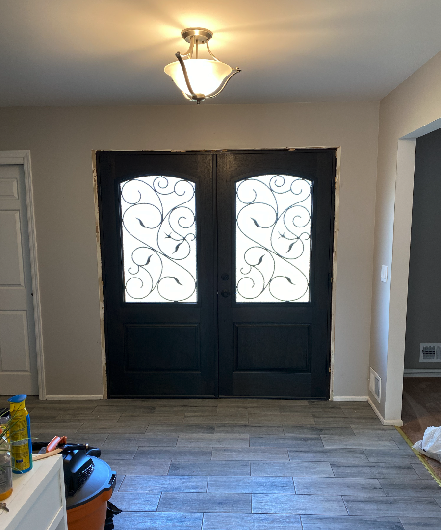 Interior view of a home entrance with a black double door with decorative wrought iron glass panels, a ceiling light fixture, light-colored walls, and wood-look flooring. Some construction or renovation work appears to be ongoing around the door fram