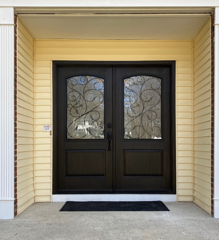 Black front door with decorative wrought iron glass panels, framed by beige siding and a black doormat on concrete porch.