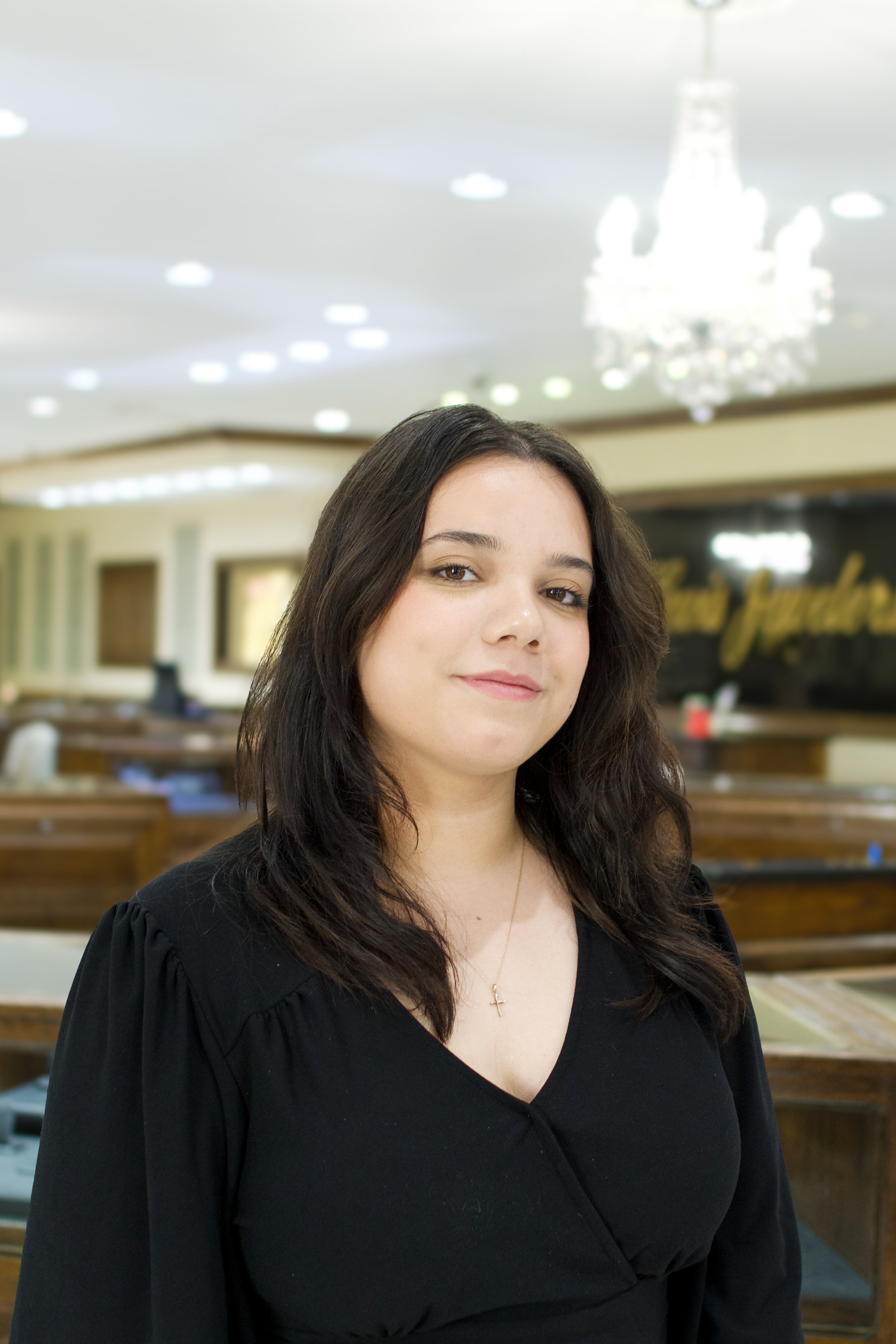 A woman with long brown hair, wearing a black top and a gold necklace with a cross, standing inside a banquet hall with a chandelier and a sign in the background.