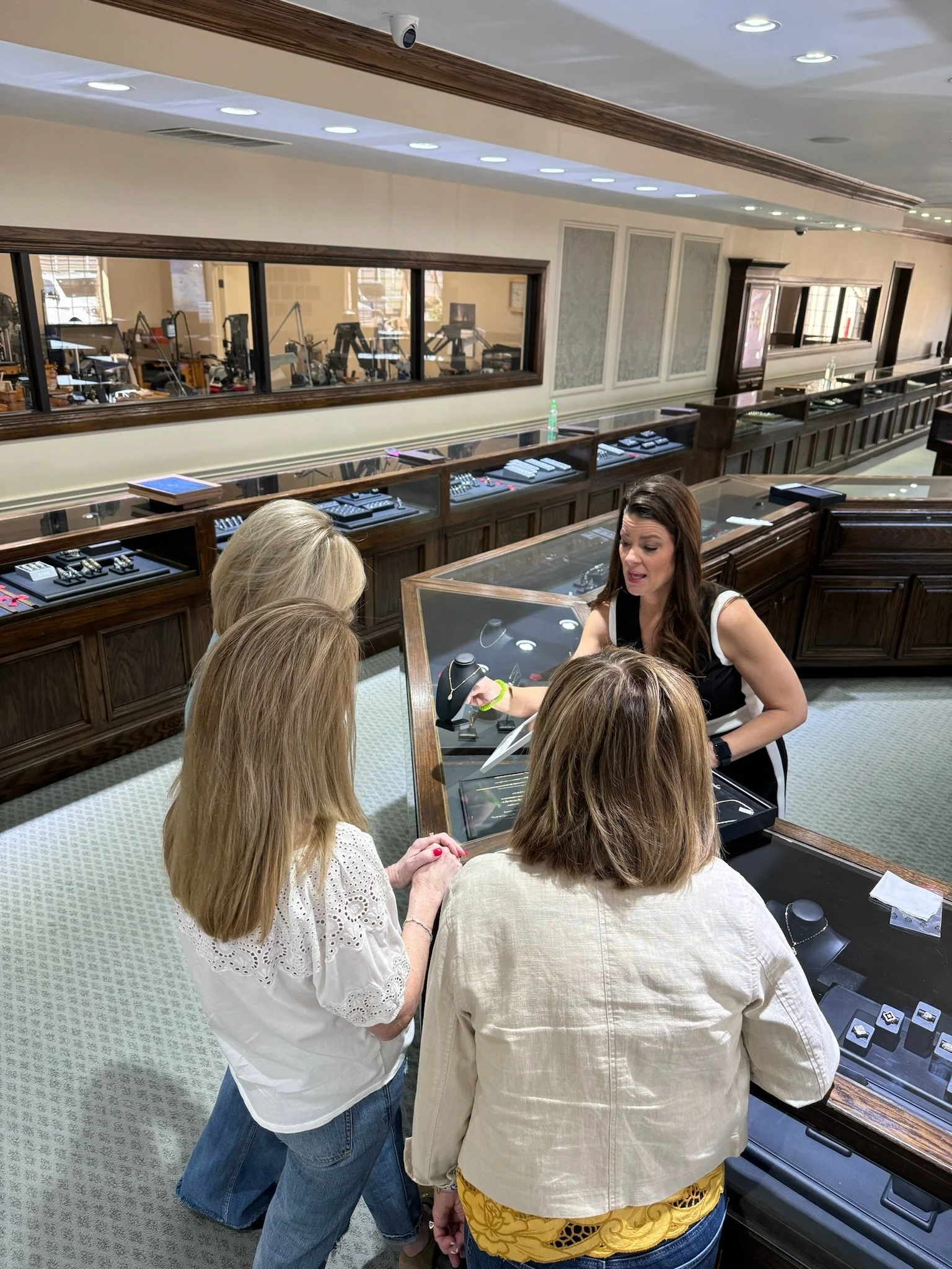 A jewelry store counter with four women looking at bracelets and necklaces under a glass display. Behind them, a woman seller is explaining. In the background, there is a window with a view into another room with fitness equipment.