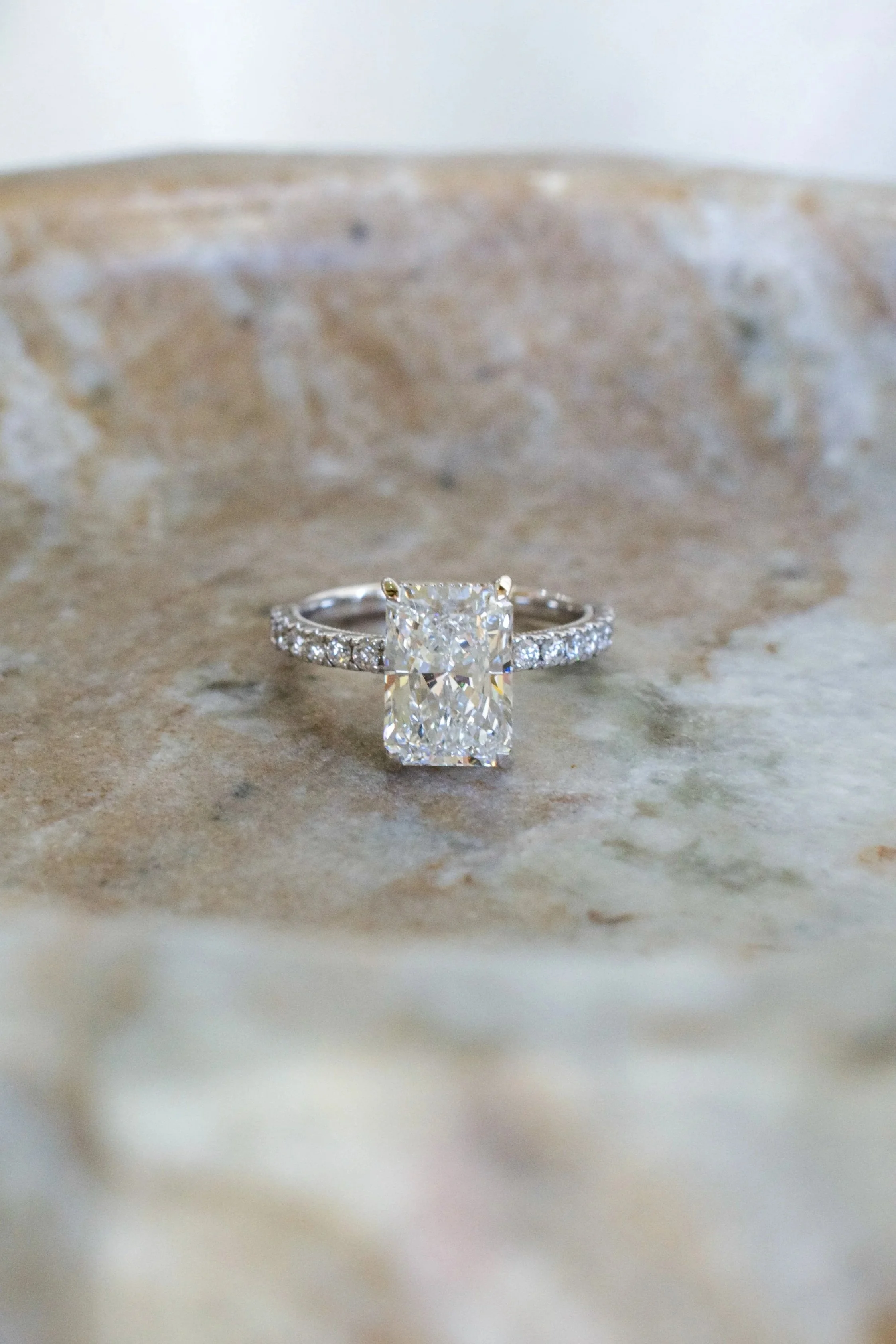 Close-up of a diamond engagement ring with a large central stone and a band decorated with smaller diamonds, placed on a textured surface.