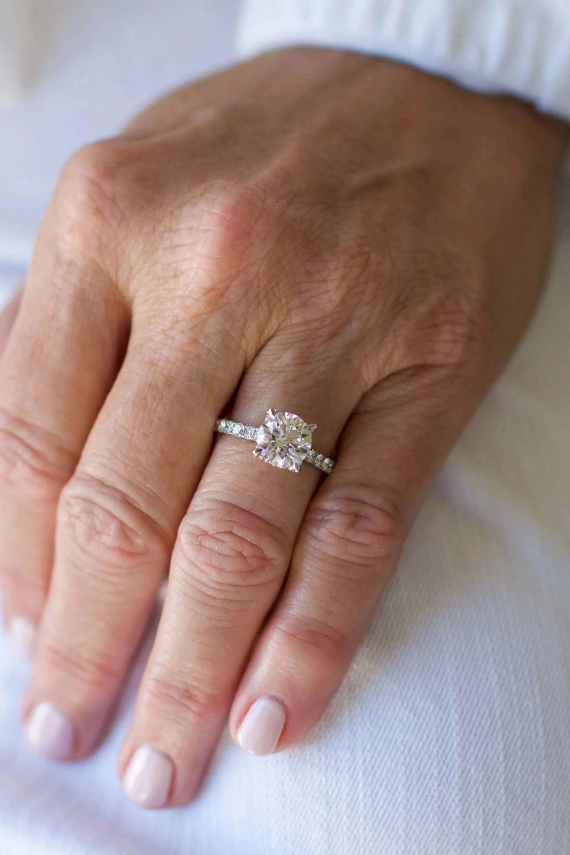 Close-up of a woman’s left hand showing an engagement ring with a large round diamond on a thin, diamond-studded band, resting on white fabric.