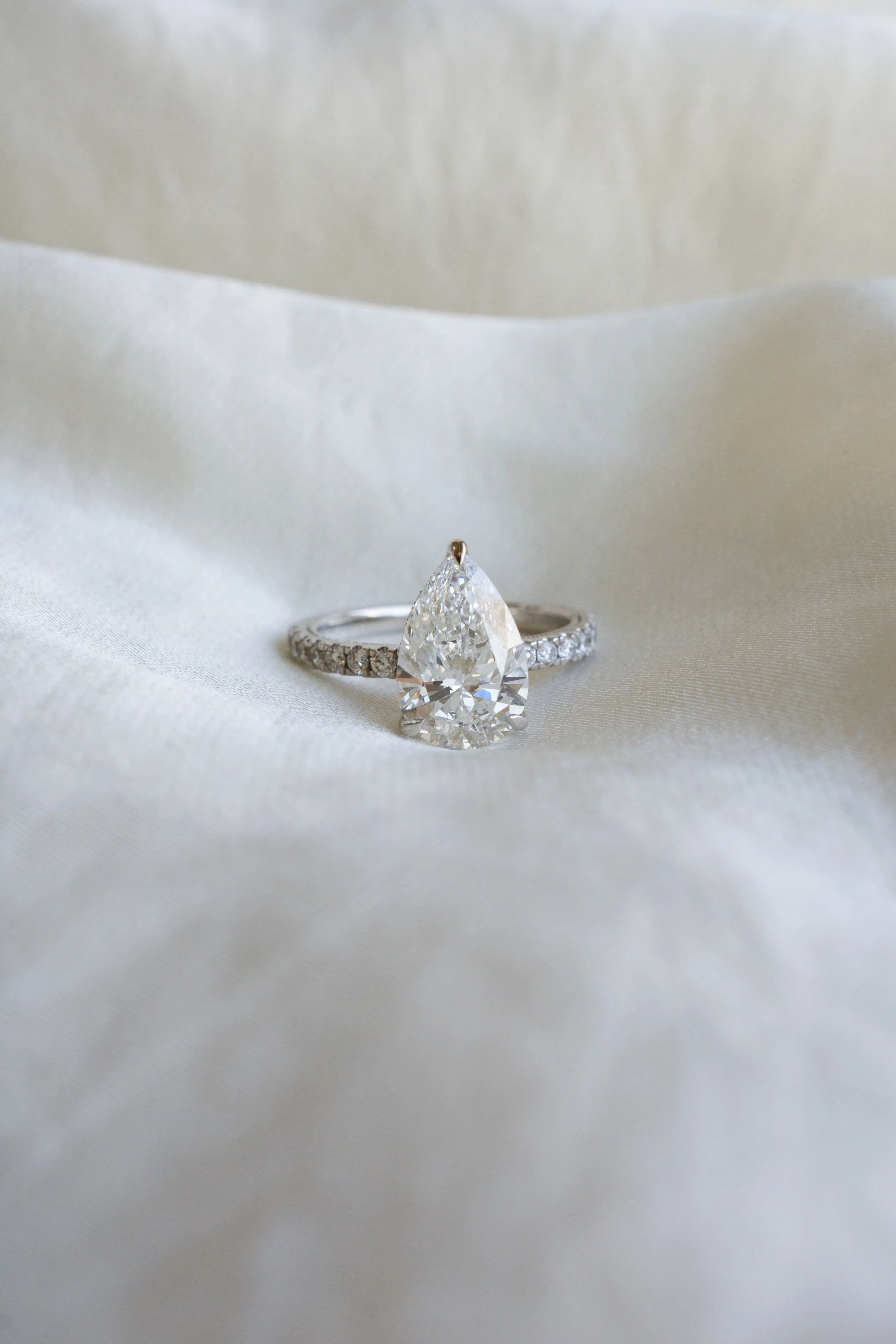 Close-up of a diamond engagement ring with a pear-shaped diamond and a band of smaller diamonds, set against white satin fabric.