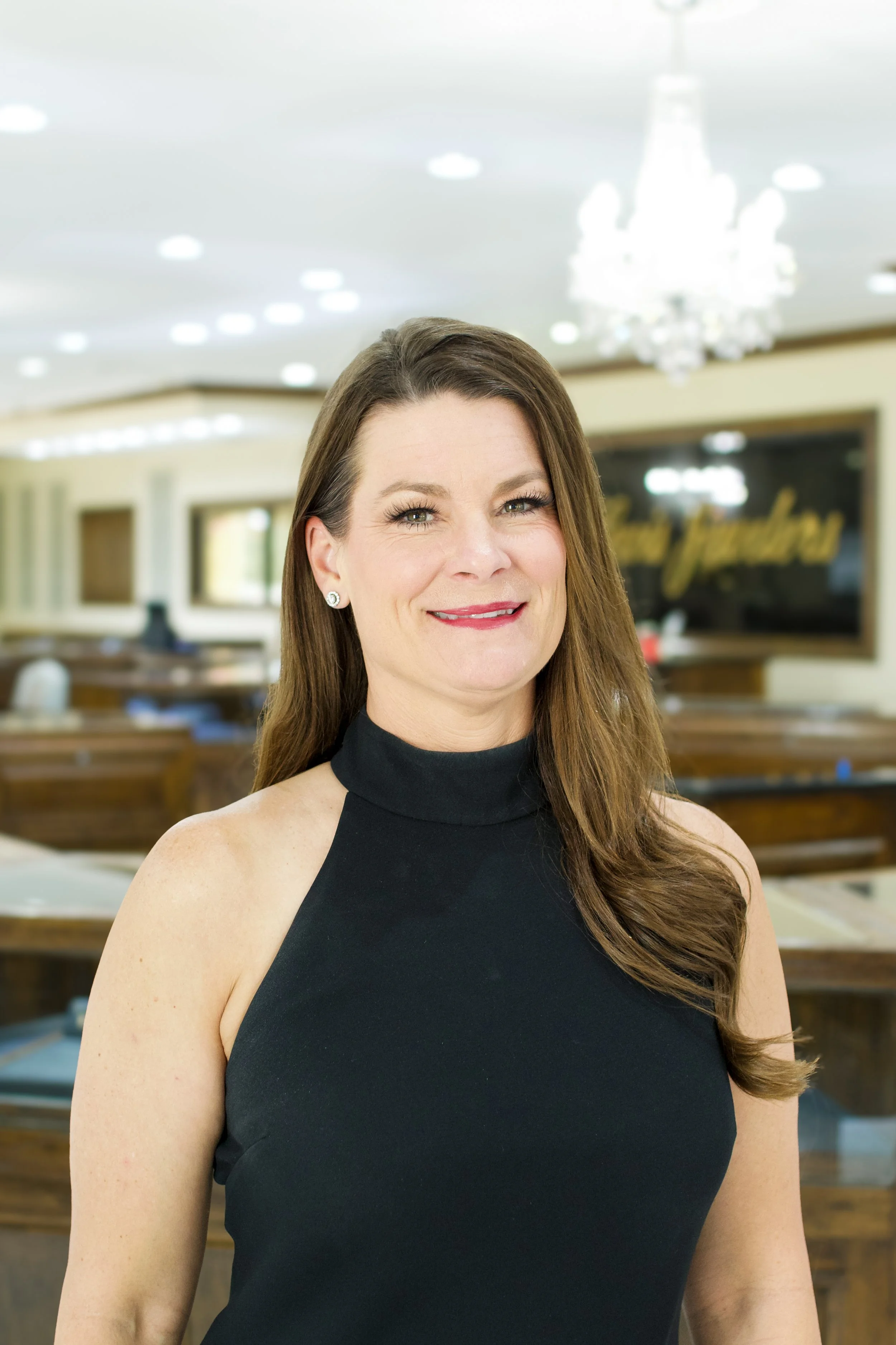 A woman with long brown hair in a black sleeveless top stands in a well-lit, elegant room with a chandelier and large windows. She is smiling at the camera.
