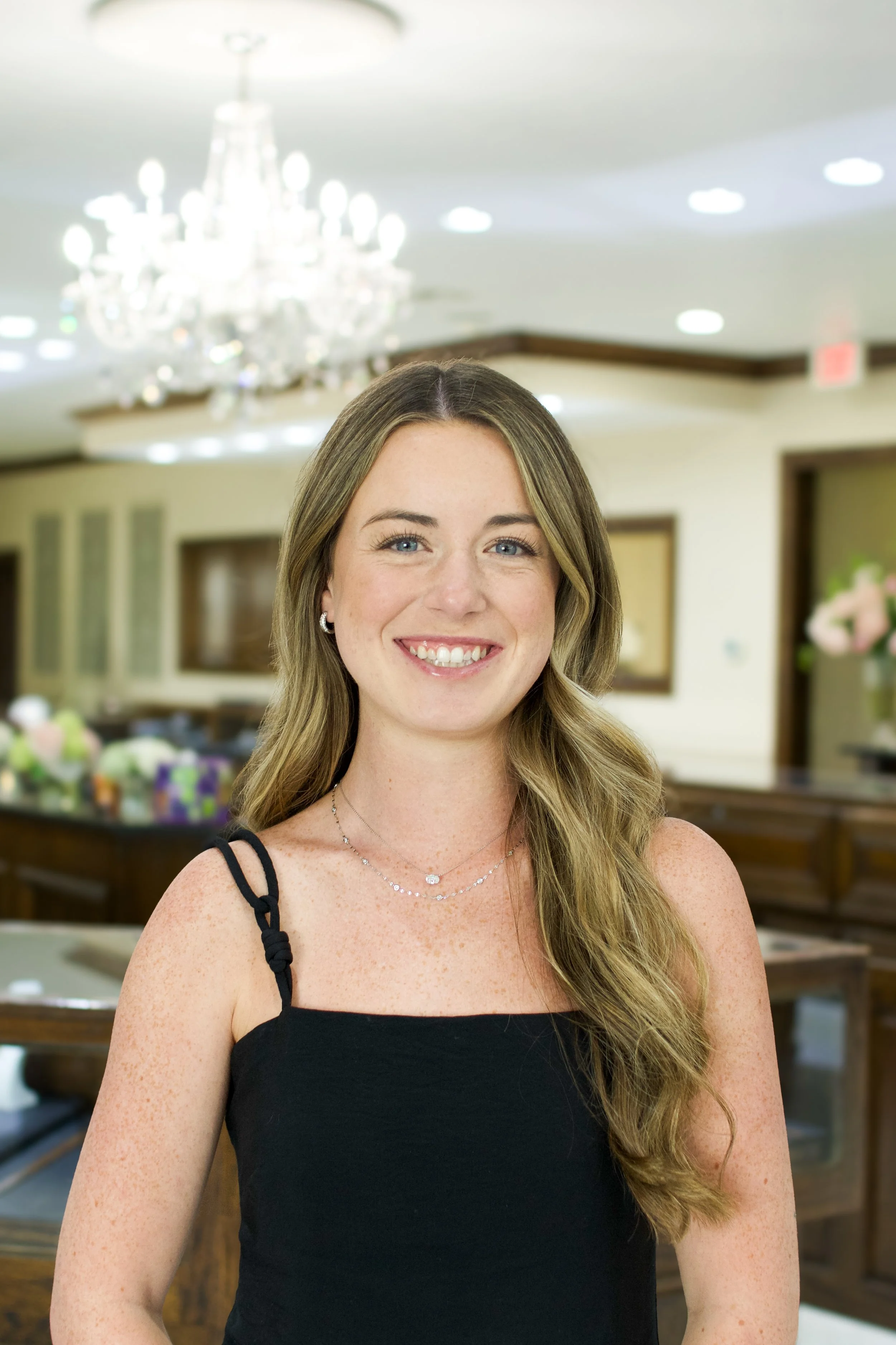 A woman with long, wavy brown hair, wearing a black sleeveless top and layered necklaces, smiling in a well-lit indoor setting with a chandelier in the background.