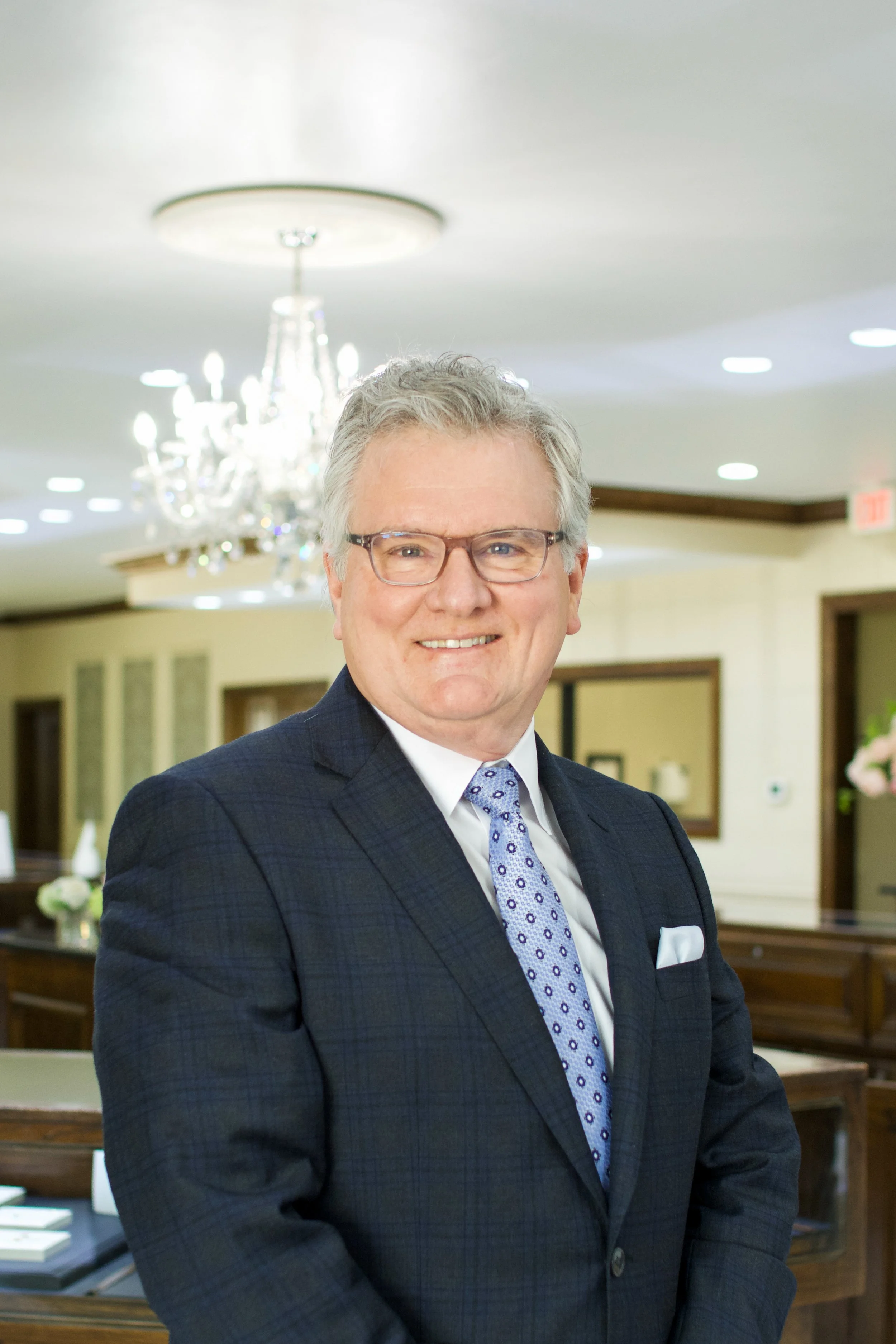 Portrait of a middle-aged man with gray hair and glasses, wearing a dark suit and a purple patterned tie, smiling in a formal setting with a chandelier and framed pictures in the background.