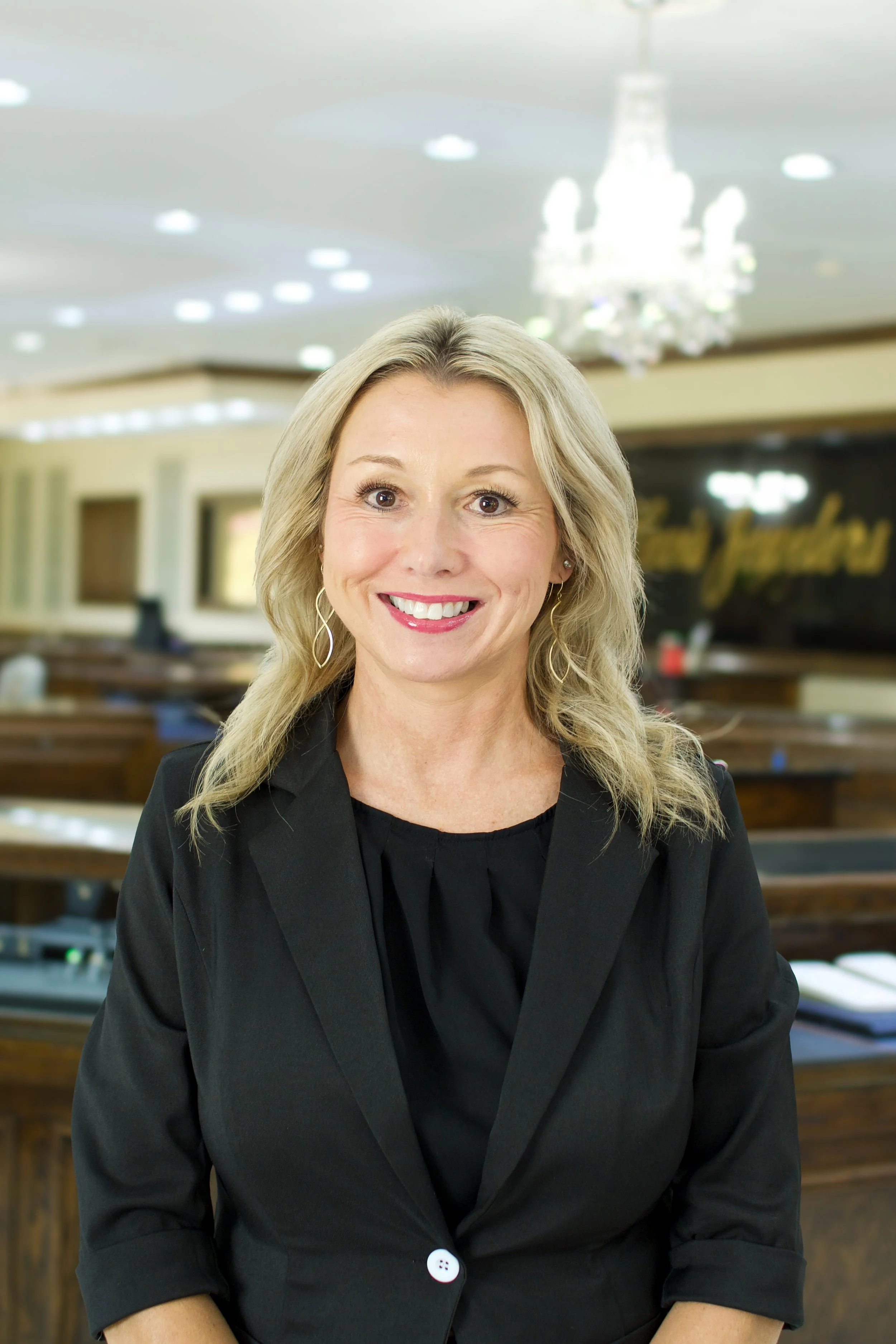 A smiling woman with blonde hair and earrings, wearing a black blazer, in a room with a chandelier and blurred background.