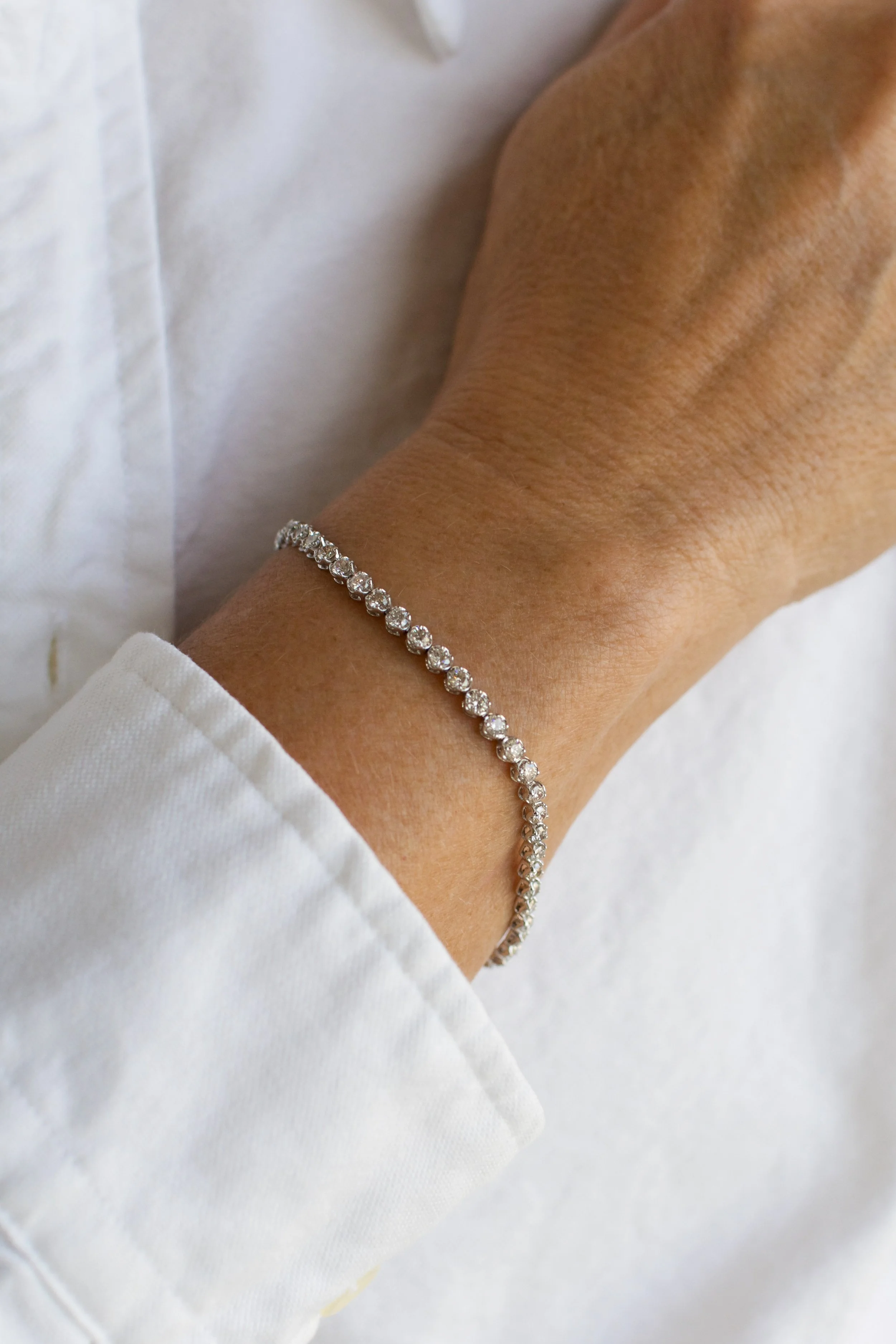 A person's wrist wearing a diamond tennis bracelet, with a white shirt and a light-colored background.