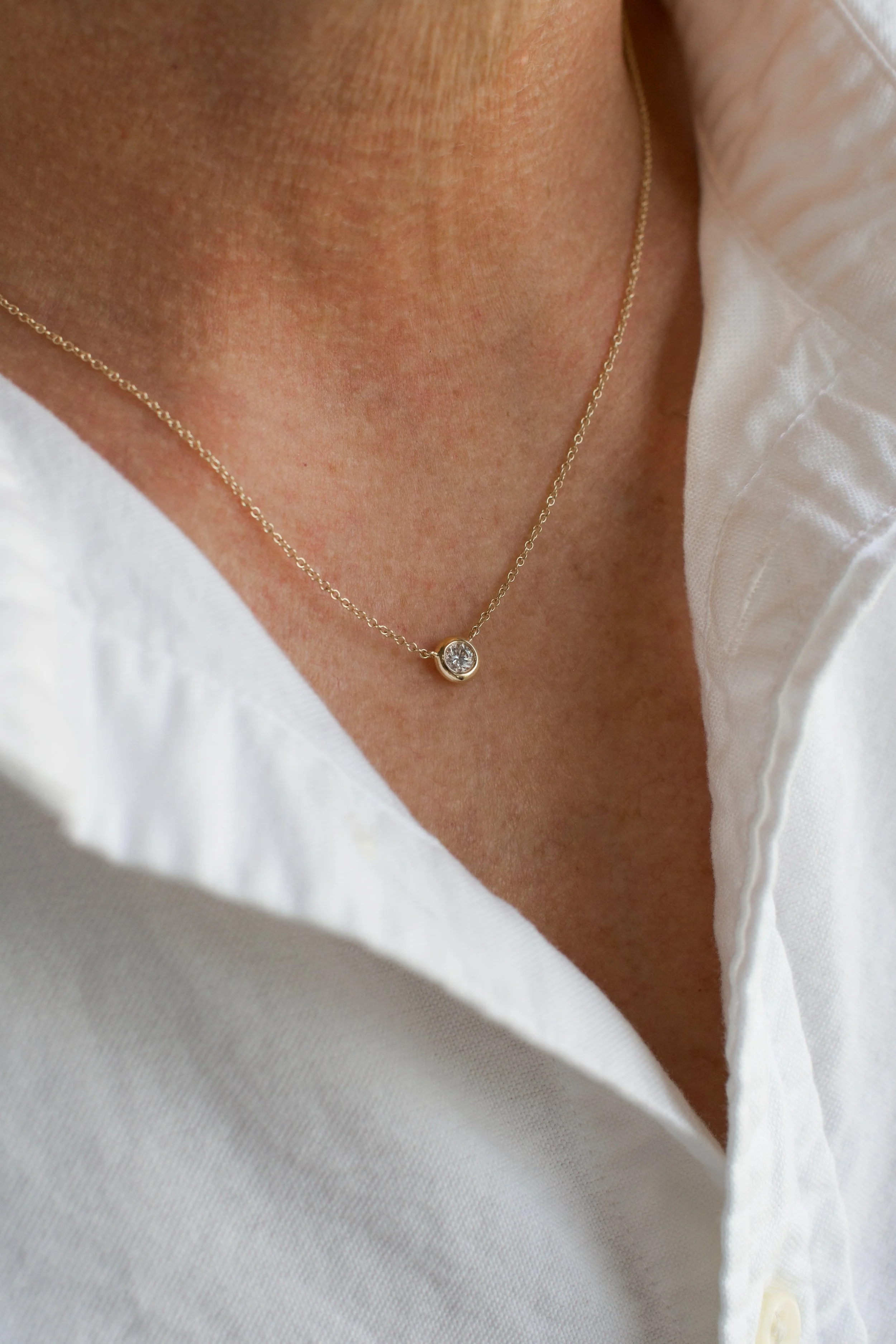 Close-up of a person's neck wearing a delicate gold necklace with a small circular pendant containing a gemstone, partially covered by a white shirt.