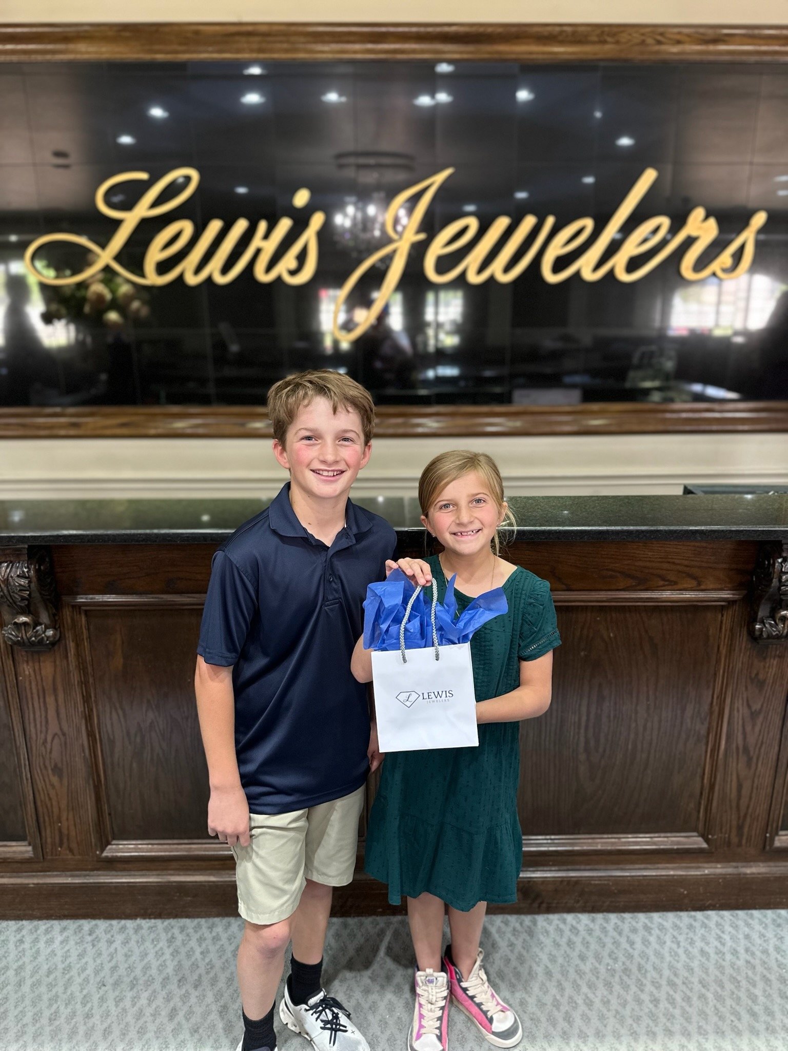 Two children, a boy and a girl, standing together in front of a Louis Jewelers sign. The boy is wearing a navy blue polo shirt, beige shorts, and white sneakers, while the girl is in a green dress with pink and white sneakers, holding a Louis Jeweler