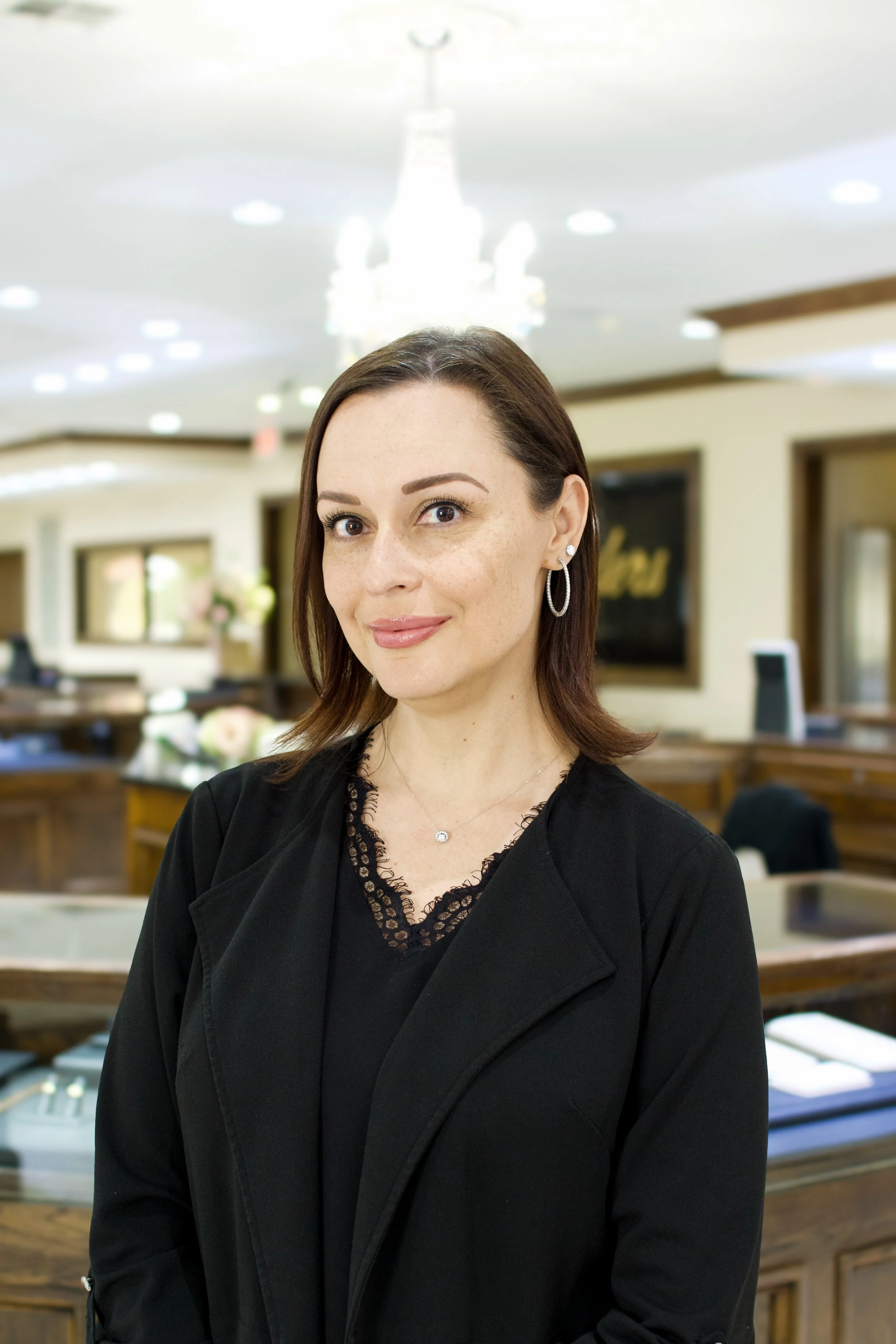 A woman with brown hair and earrings, wearing a black blazer over a black lace top, standing inside a room with a chandelier, wooden furniture, and decorative items in the background.