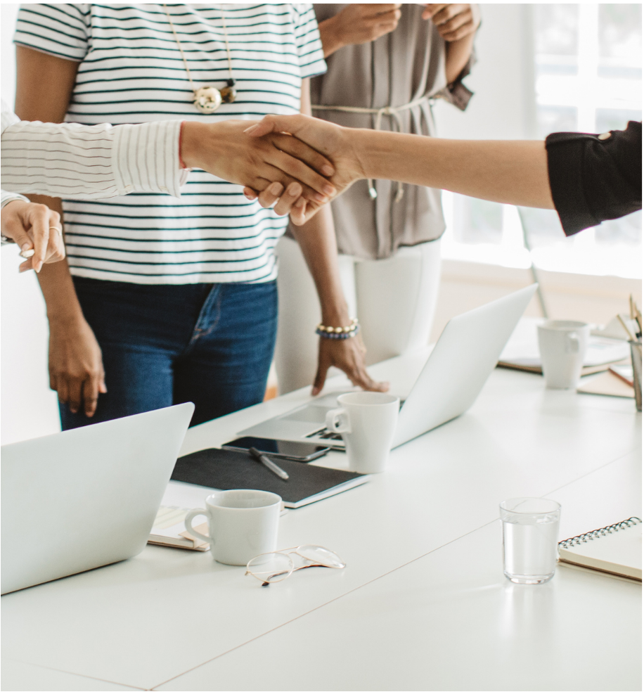 People shaking hands over a table with laptops, notebooks, coffee cups, a glass of water, and glasses on it in a bright office setting.