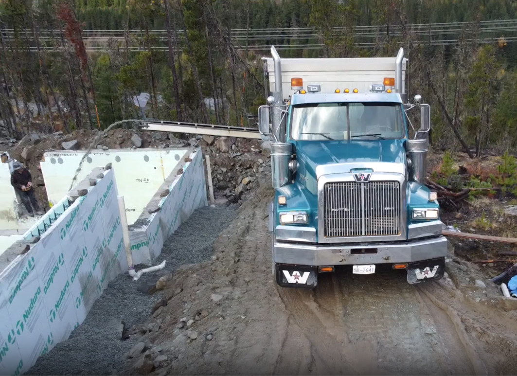 A construction site with a large blue stone slinger truck on a dirt road, adjacent to a partially built concrete structure with insulation panels, surrounded by forest and power lines in Whistler, BC.