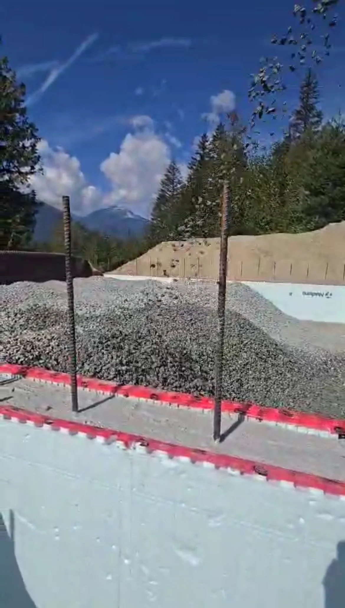 Construction site with a large pile of gravel, steel rebar sticking out of the gravel, and mountainous background with trees, blue sky, and clouds.
