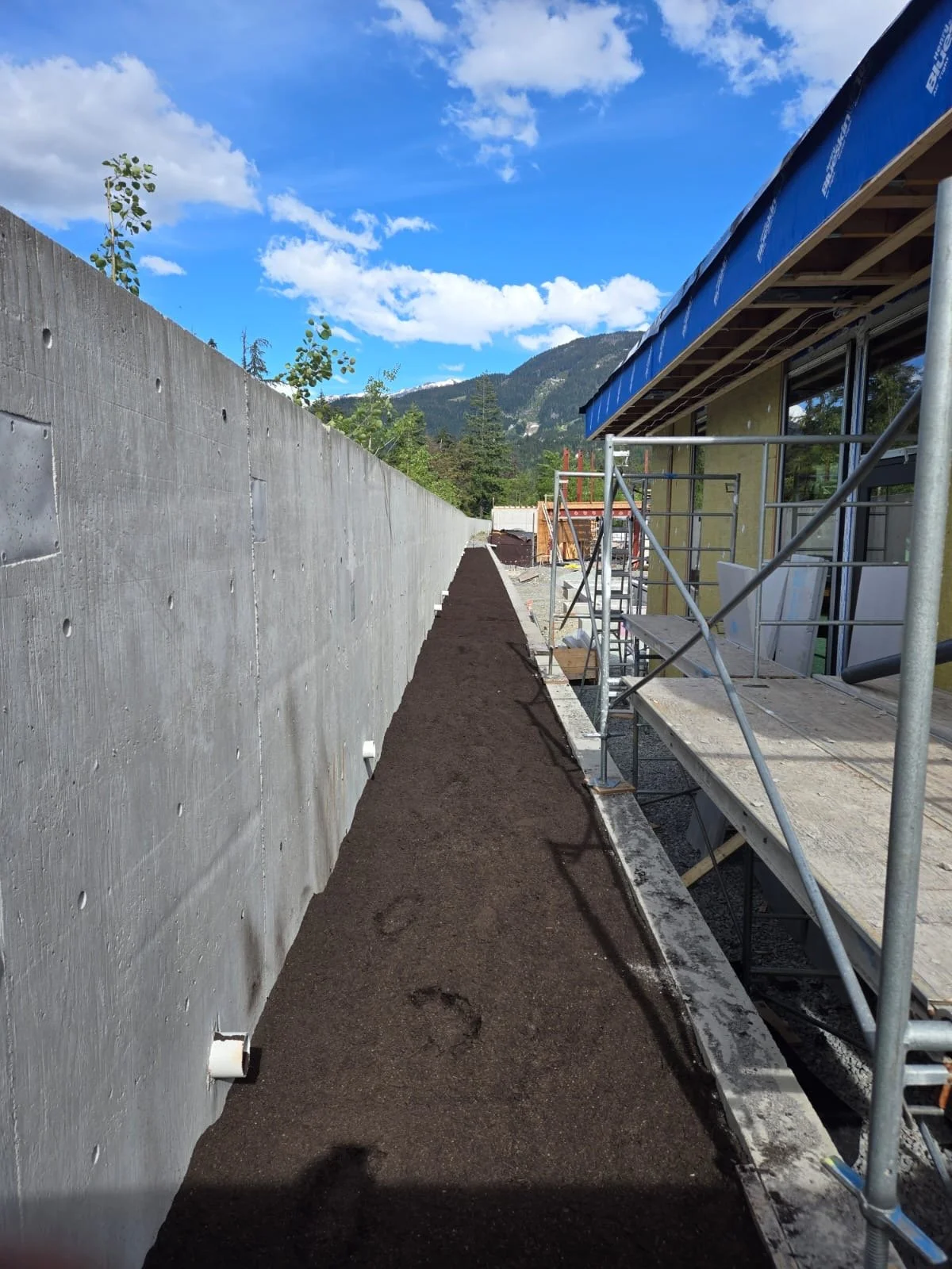 Freshly placed soil on a residential site, house with scaffolding, mountains and a partly cloudy sky in the background.