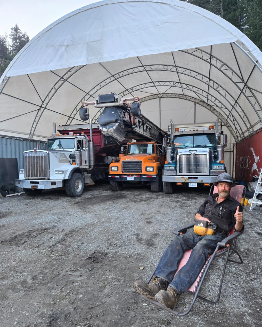 Drew sitting in a reclining chair in front of a large, open-sided storage tent, giving a thumbs-up. Behind him are his three large trucks and construction equipment parked under the tent.