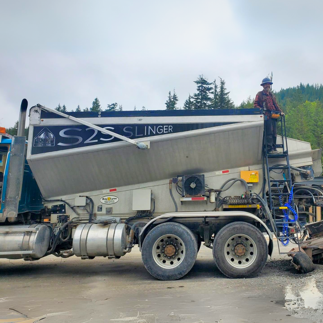 Drew standing on a platform with his stone slinger beside construction site with Sea-to-Sky mountains and trees in the background.