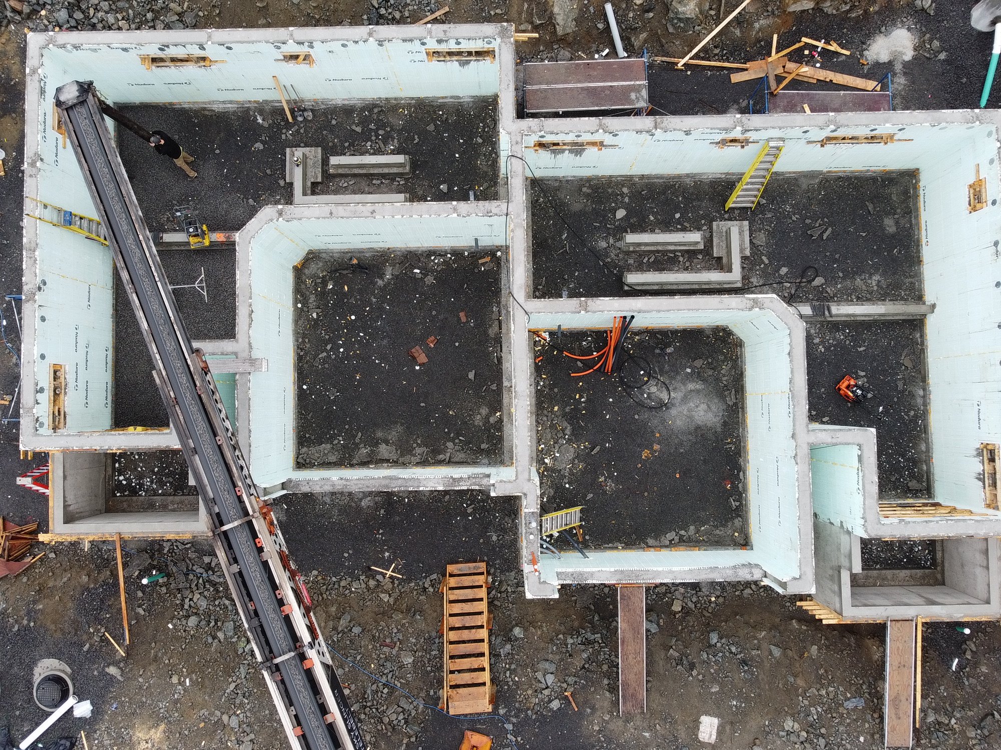 Aerial view of a construction site showing the framing of a building with concrete walls, construction equipment, and the conveyor truck feeding in backfill material.