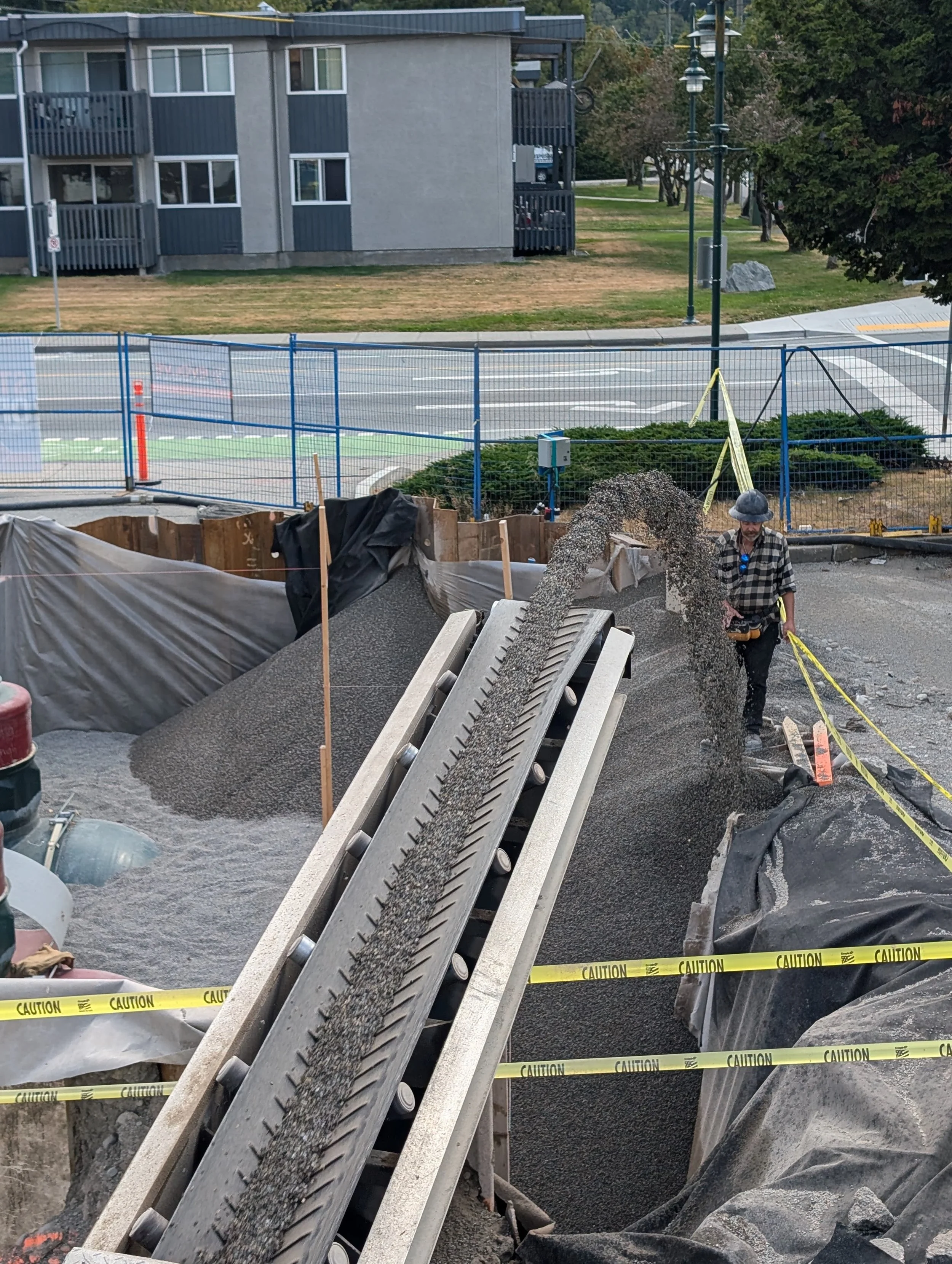 Delivering backfill material to a construction site with a conveyor belt, caution tape, and construction materials, in an urban setting.