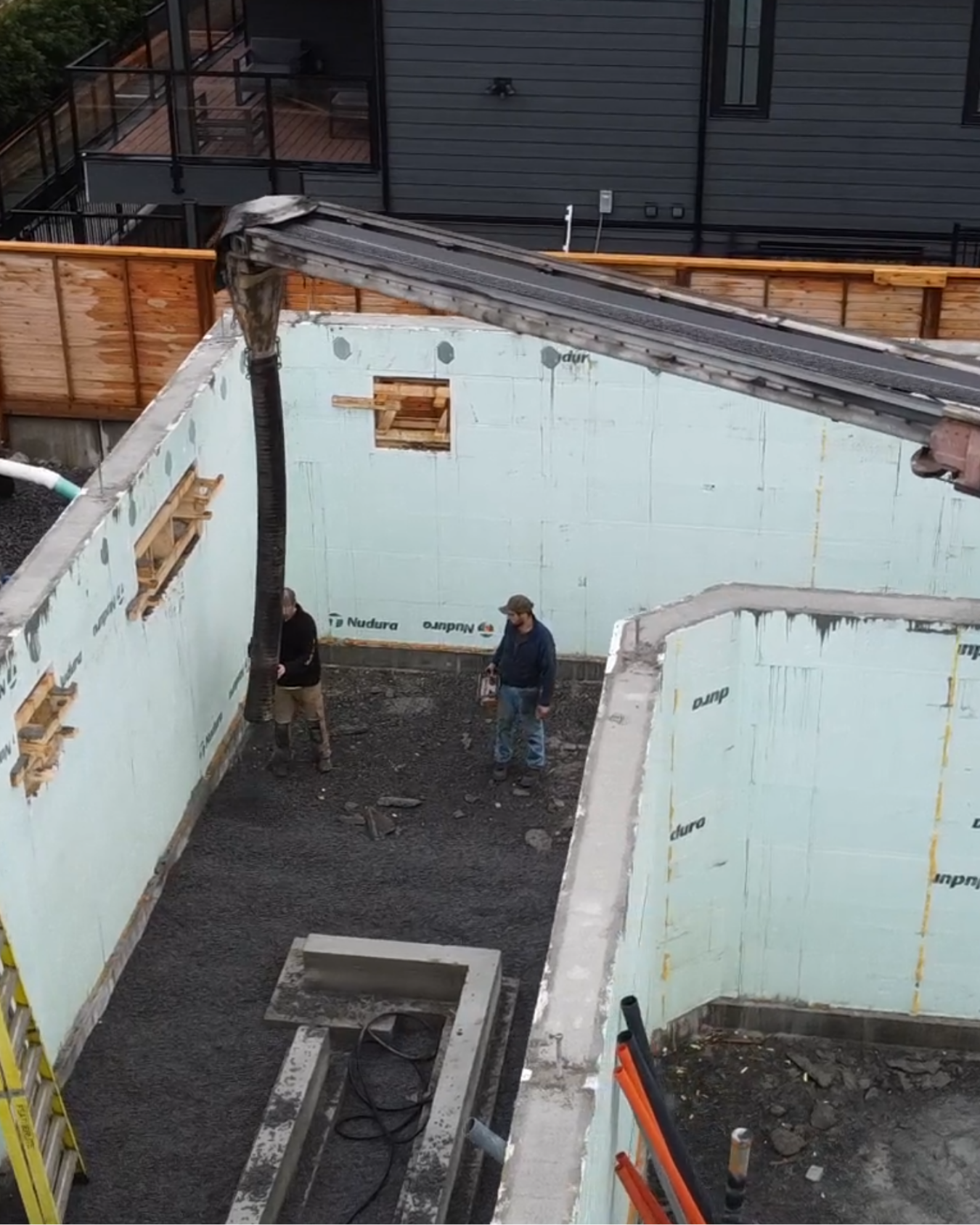 Construction site with two workers inside a foundation area placing material, walls lined with insulation, and building materials scattered around.