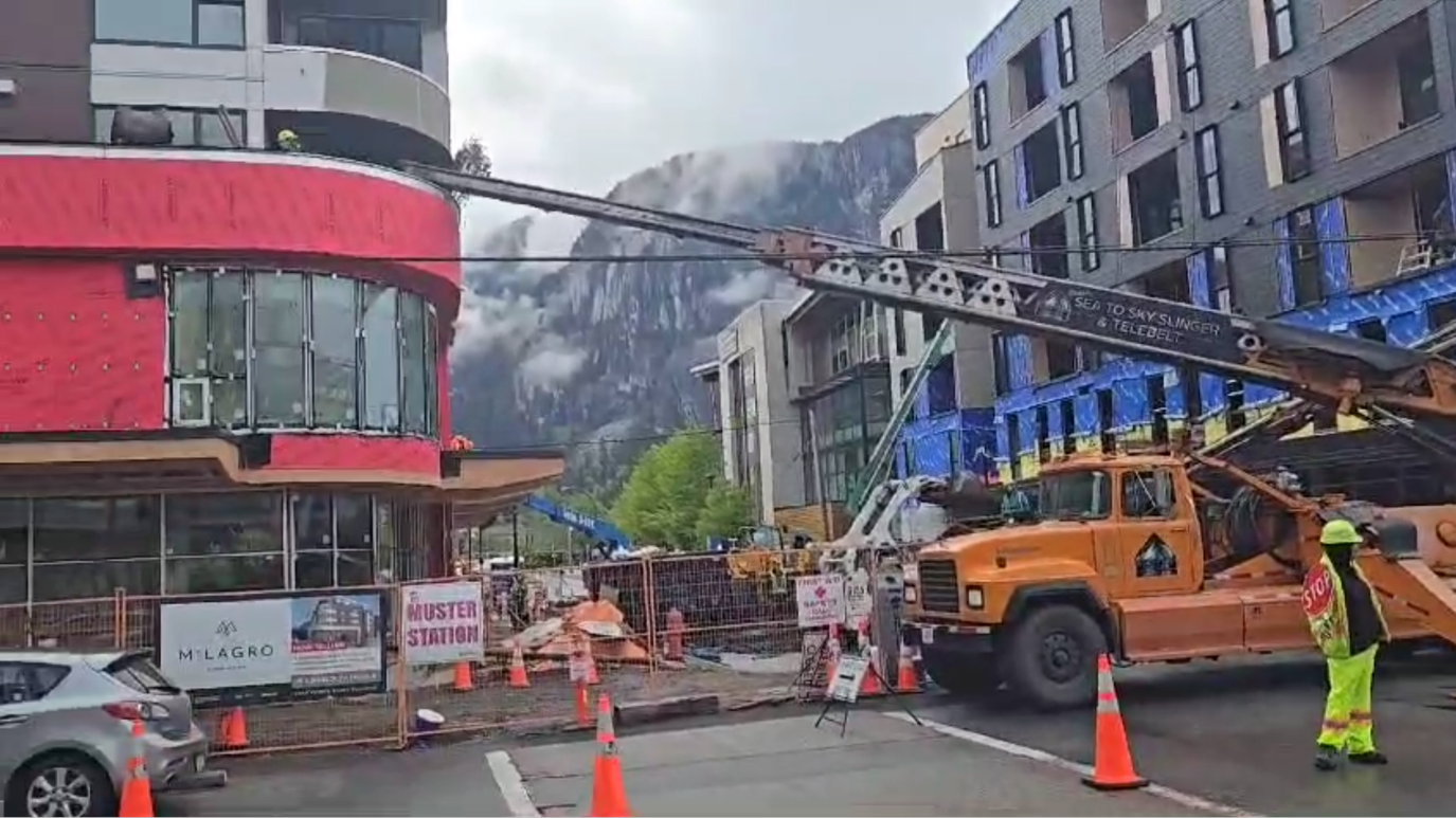 Construction site on a street in Squamish with a large conveyor on an orange truck delivering soil to a second-storey garden bed. Construction barriers, and a worker in a safety vest and helmet. Squamish chief mountain is visible in the background.