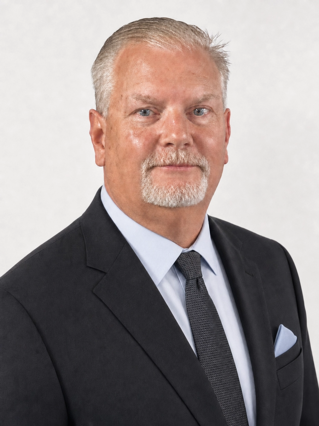 Headshot of a middle-aged man with short gray hair, beard, and blue eyes, wearing a dark suit, light blue dress shirt, and a dark tie, against a plain white background.