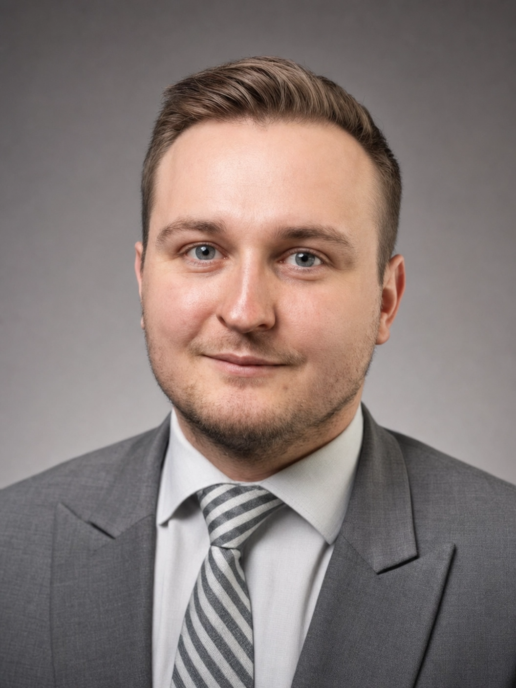 Professional headshot of a young man with light brown hair and blue eyes, wearing a gray suit, white shirt, and striped tie, against a gray background.