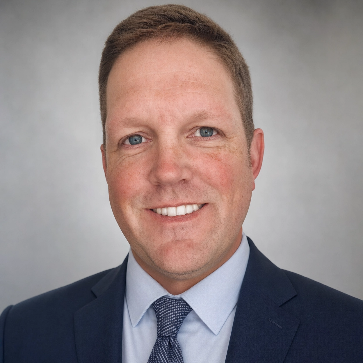 A professional headshot of a man with short light brown hair, blue eyes, and fair skin, dressed in a navy blue suit, light blue shirt, and patterned tie, smiling against a neutral gray background.