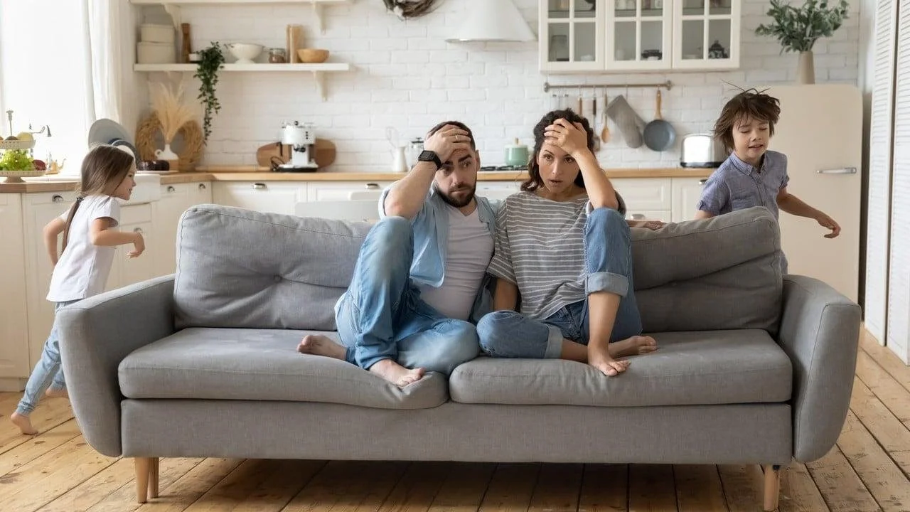 A worried couple sitting on a gray sofa in a kitchen, with two children playing nearby.