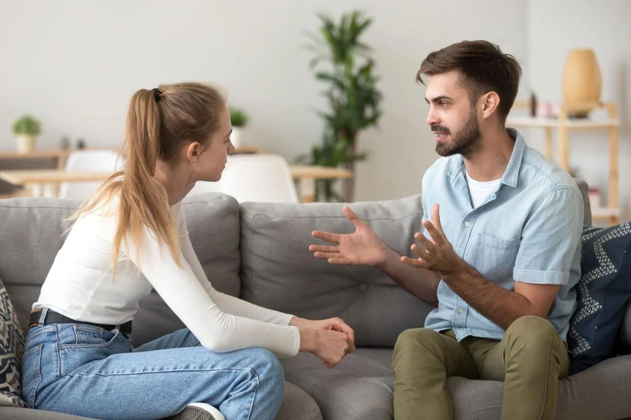 A young woman and a young man are sitting on a gray couch having an intense conversation. The woman appears to be listening, while the man is gesturing with his hands, looking frustrated or upset. In the background, there are plants and home decor items.