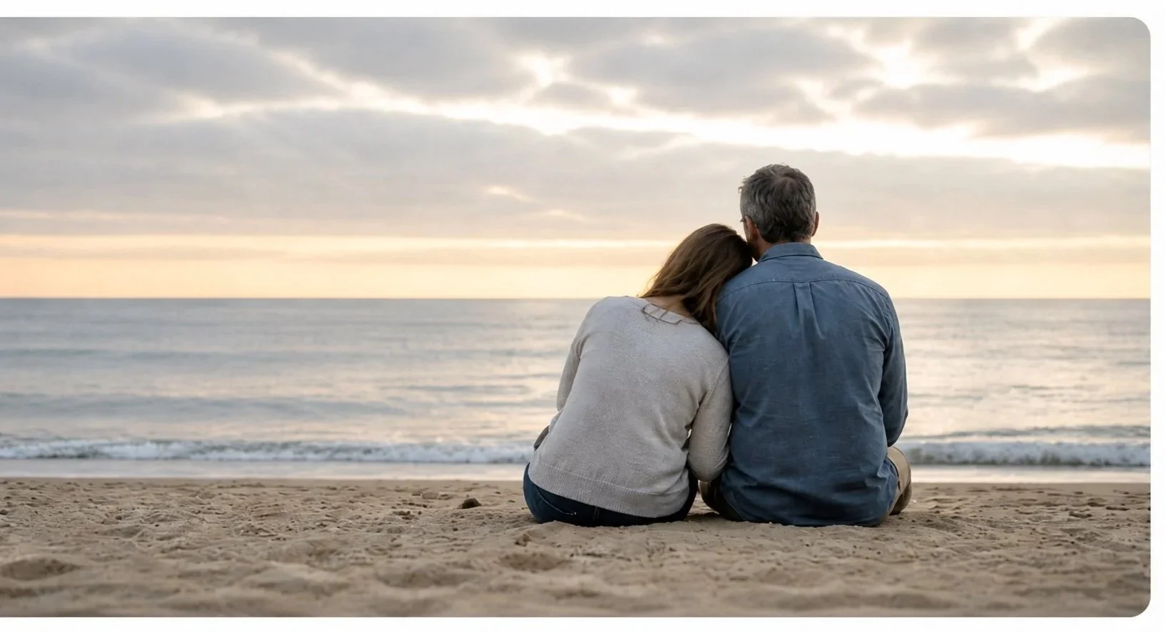 A couple sitting on the beach, facing the ocean, with the woman resting her head on the man's shoulder during sunset.