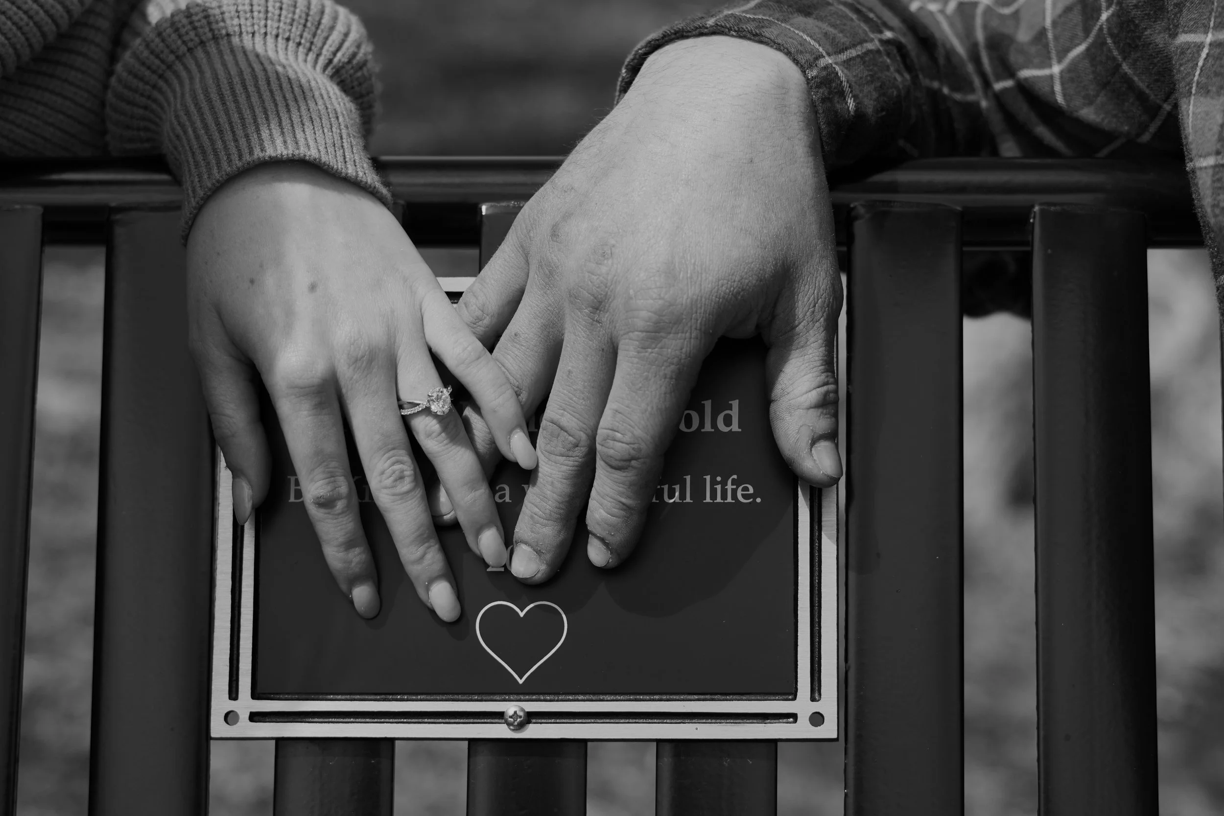 Close-up of a person's hands resting on a park bench, with the left hand wearing an engagement ring. The hands are on a sign that says "Hold beautiful life" with a heart symbol. vintage style photography northern nj