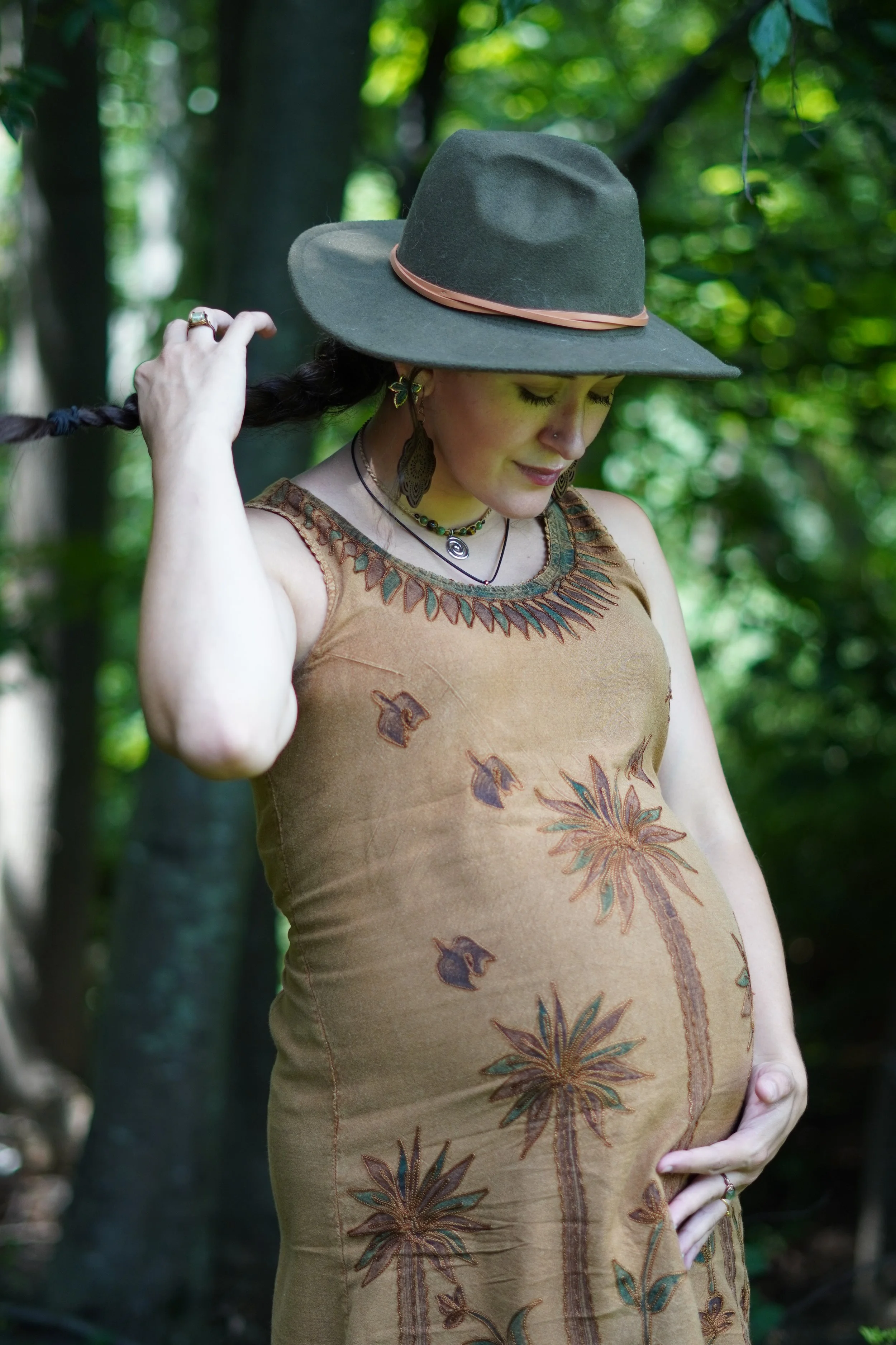 A woman with curly hair wearing a wide-brimmed hat, a sleeveless dress with a floral and leaf pattern, and jewelry, standing outdoors in a forest. Maternity photographer