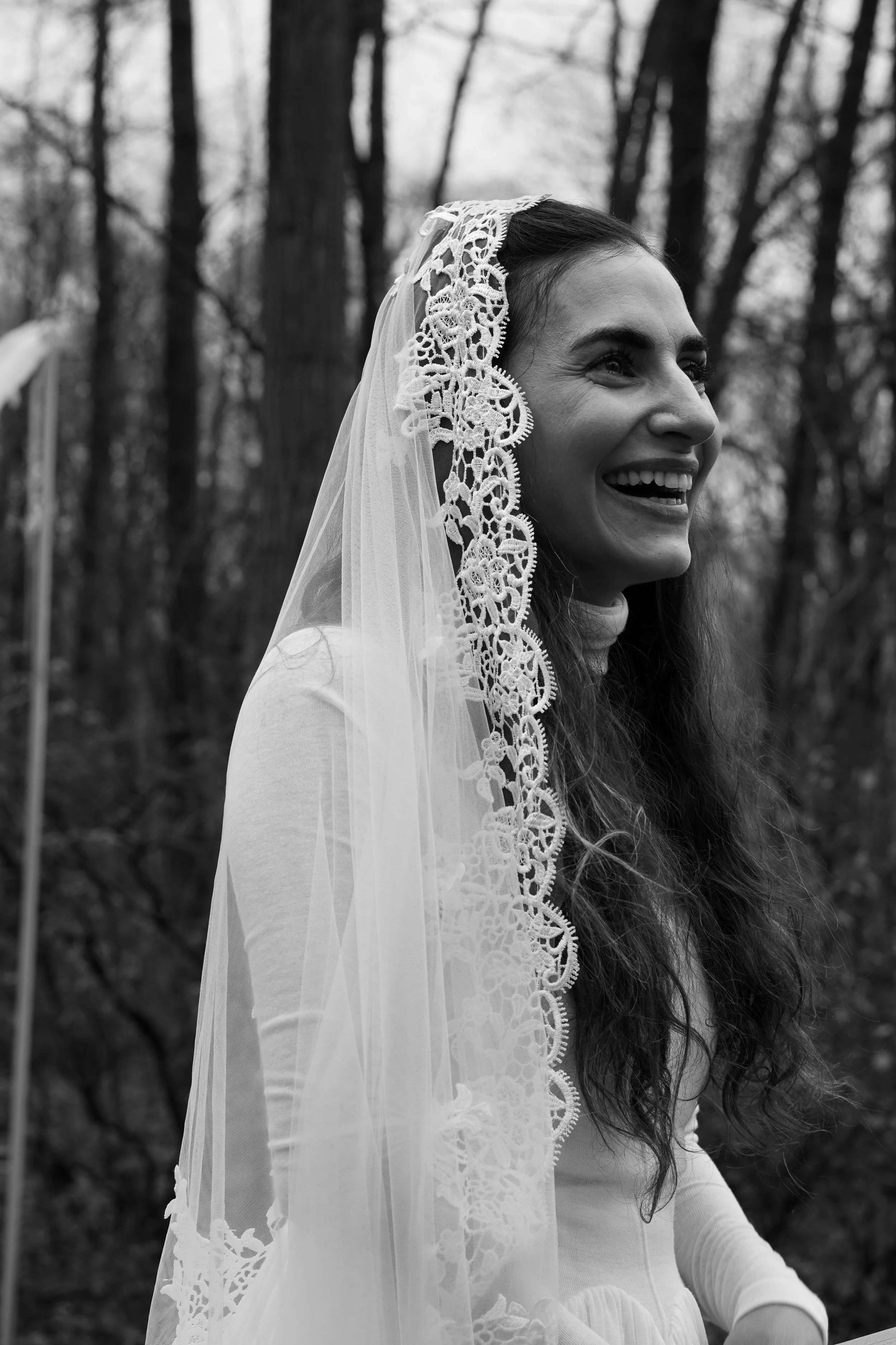 A woman wearing a lace-trimmed veil and a light-colored dress, smiling outdoors with trees in the background. Cheap wedding photographer nj