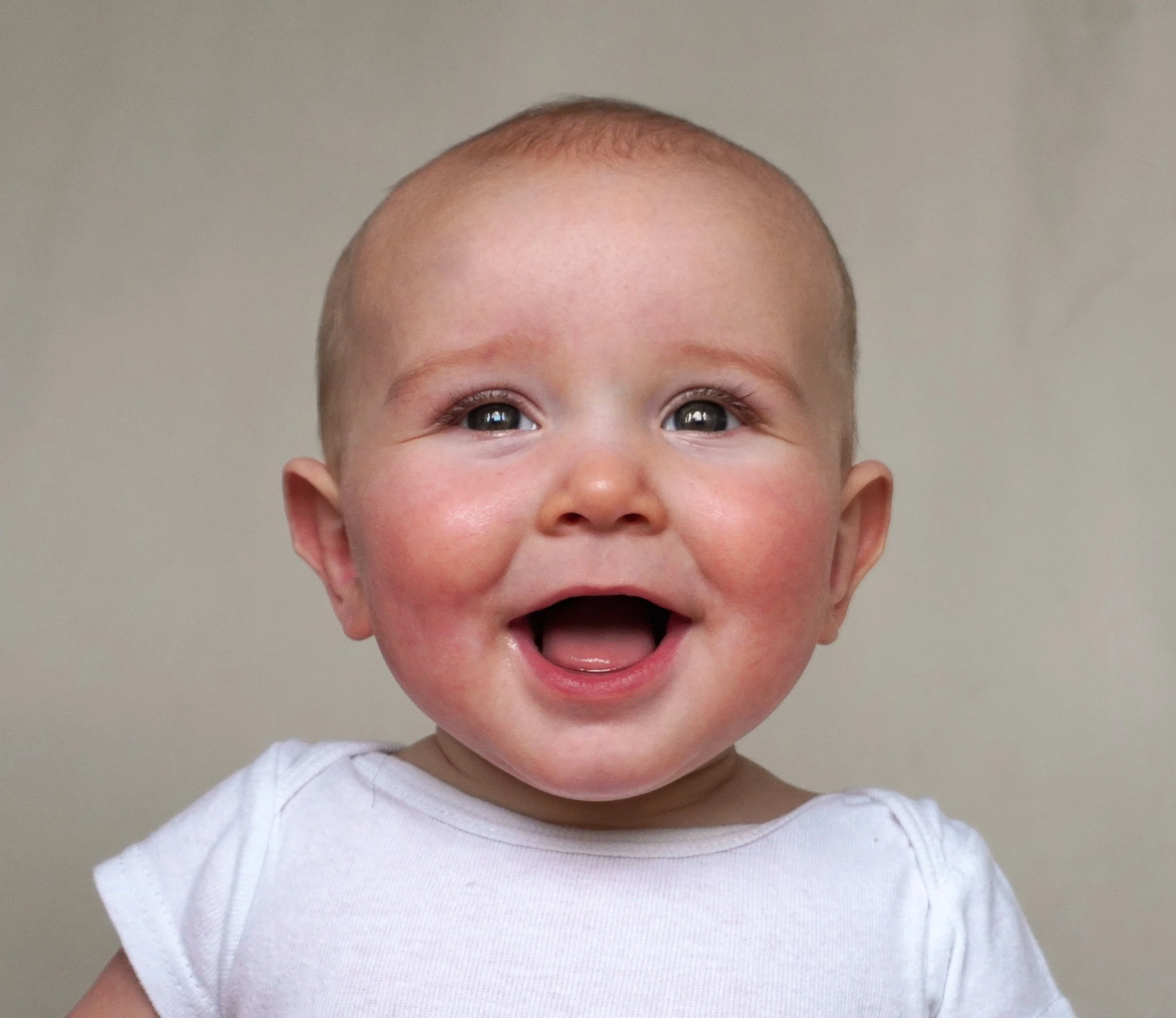 A smiling baby with a bald head, wearing a white shirt, against a plain background. Baby photography northern nj