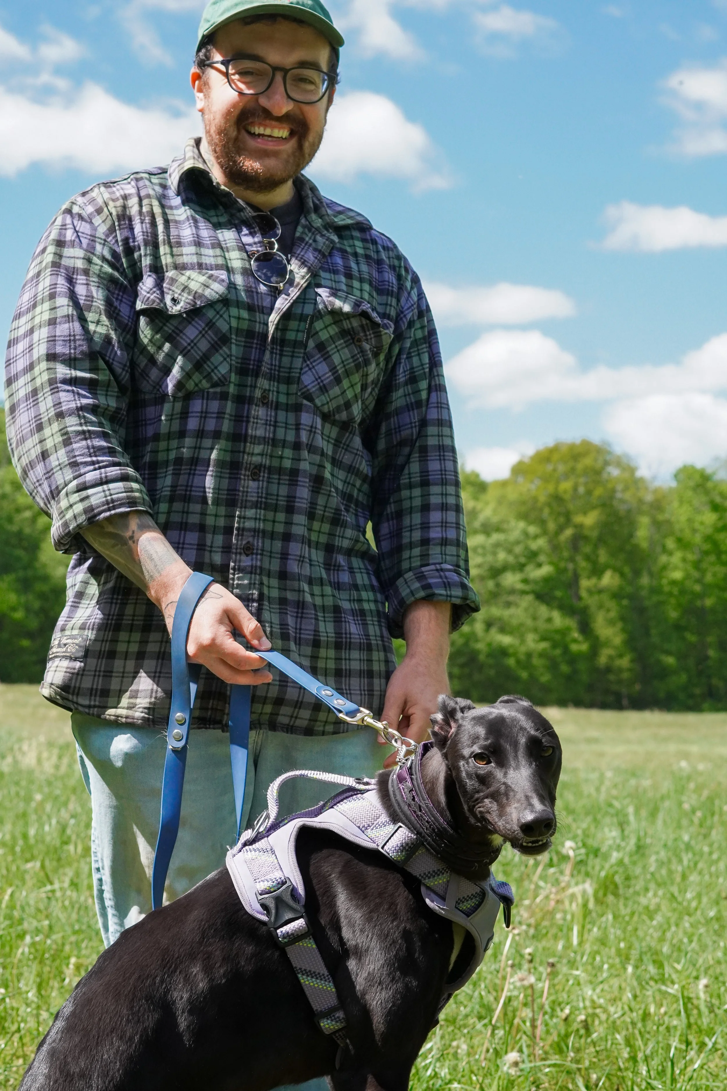 A man with glasses, a beard, and a green cap, smiling while holding a leash attached to a black dog wearing a harness, standing in a grassy field with trees and a partly cloudy sky in the background. Dog photographer northern nj